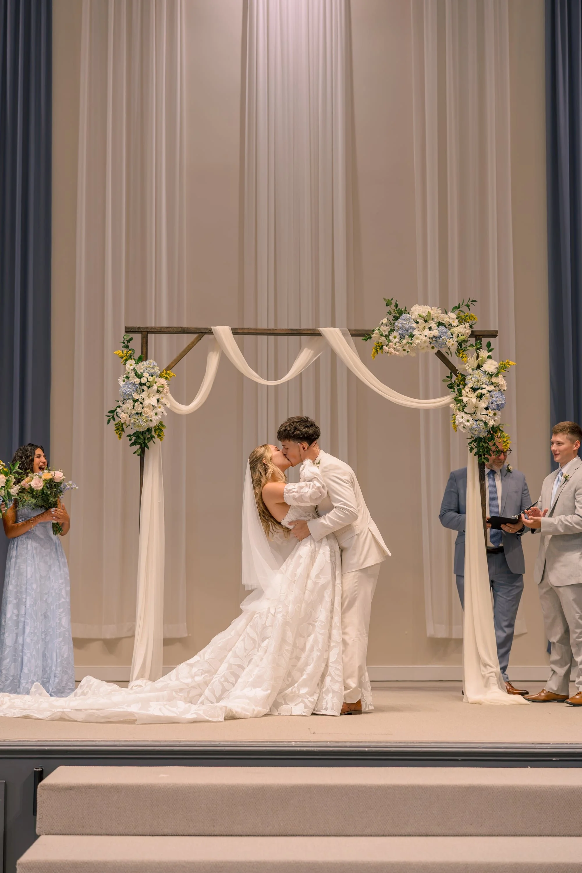 A bride and groom kissing under a floral wedding arch during a ceremony with wedding guests and an officiant in the background.