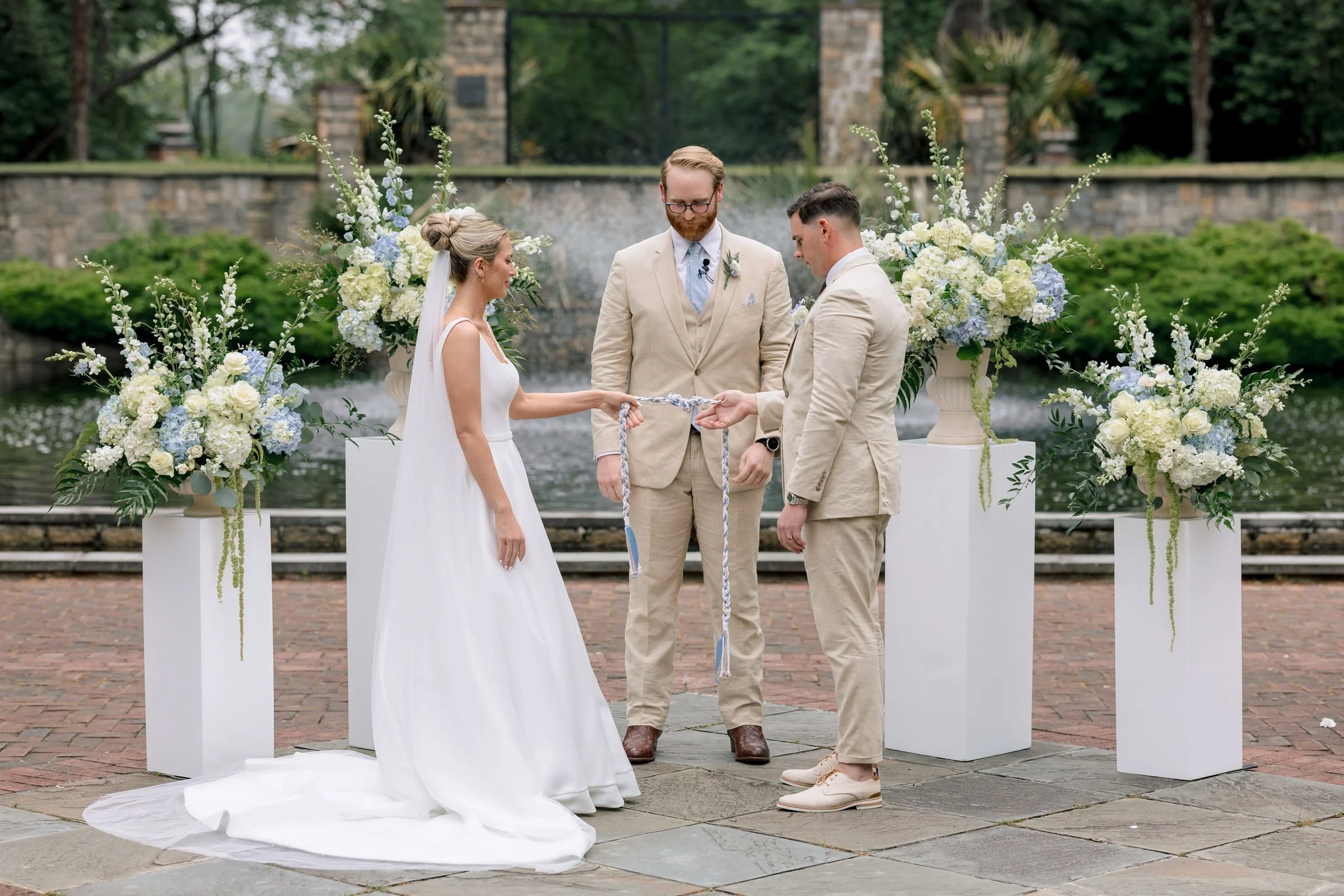 A wedding ceremony outdoors with a bride, groom, and officiant. The bride wears a white dress with a veil, and the groom and officiant wear beige suits. They stand in front of floral arrangements with white and light blue flowers, near a fountain and