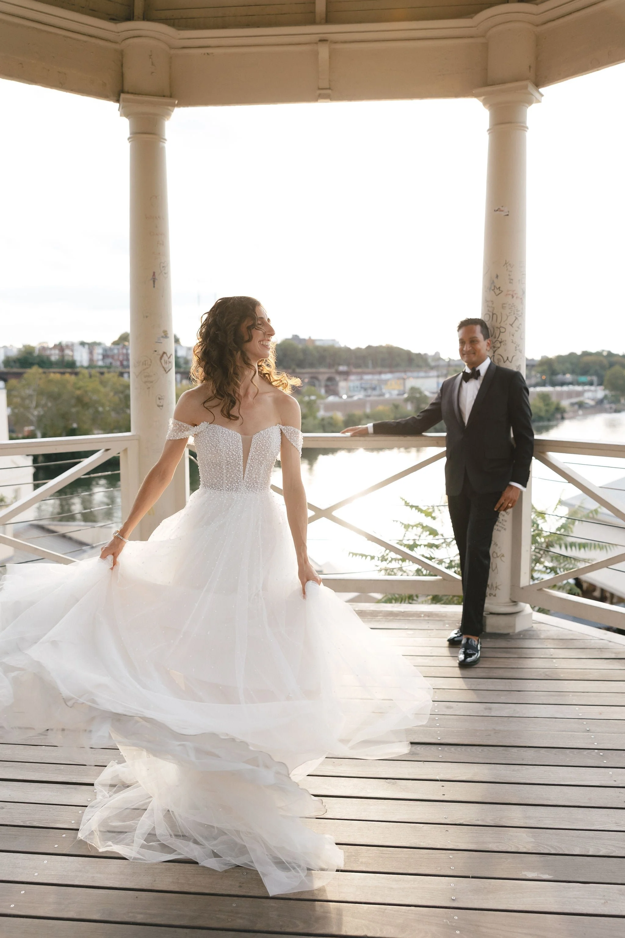 A bride in a white wedding gown with off-the-shoulder sleeves and a full skirt, smiling and holding her dress, standing on a wooden deck. A groom in a black tuxedo with a bow tie stands nearby, smiling and extending his hand. They are under a gazebo 