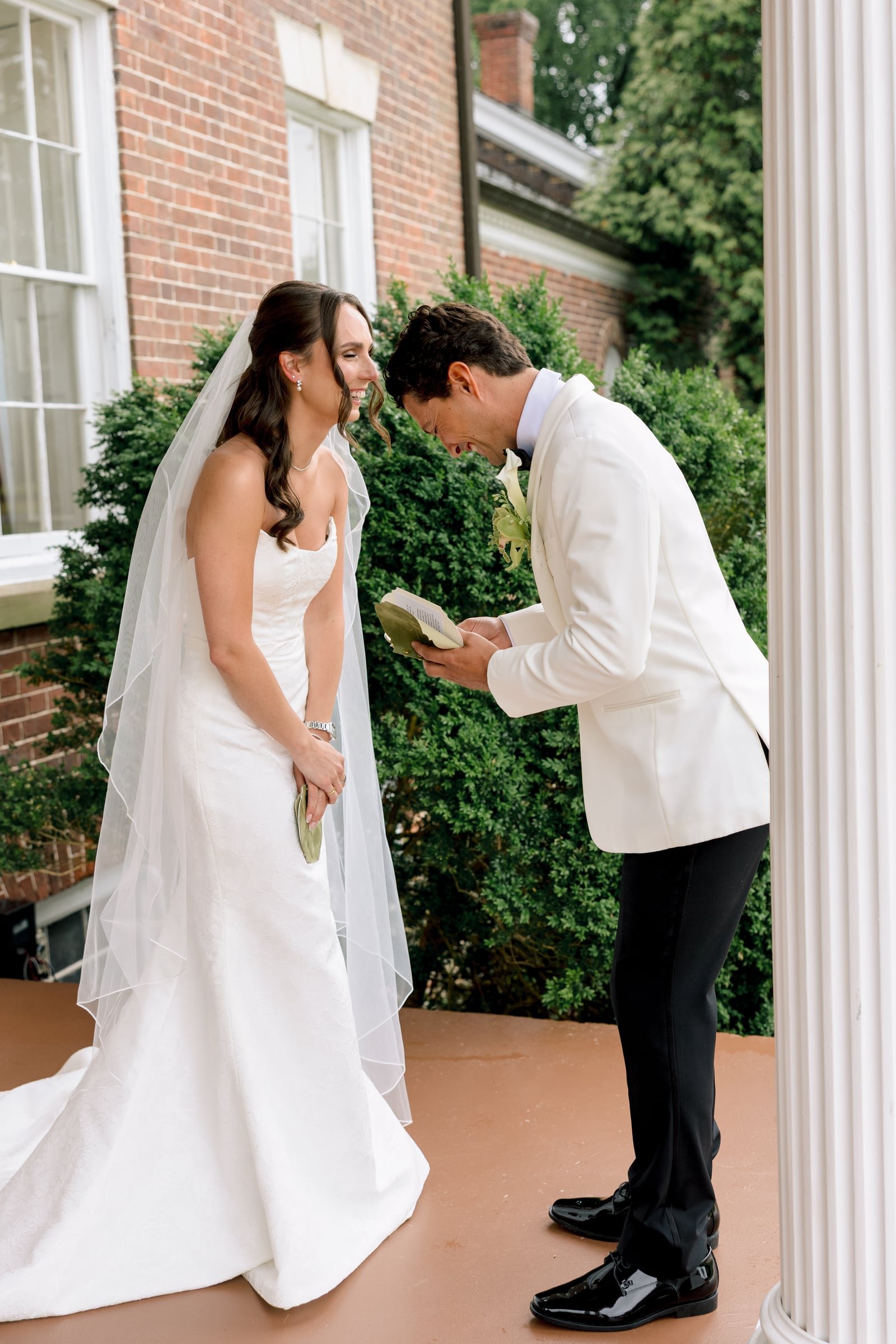 A bride and groom standing on a porch, the groom laughing and looking down at a book or note, the bride smiling and looking at the groom, during a wedding ceremony outdoors with green bushes and a brick house in the background.