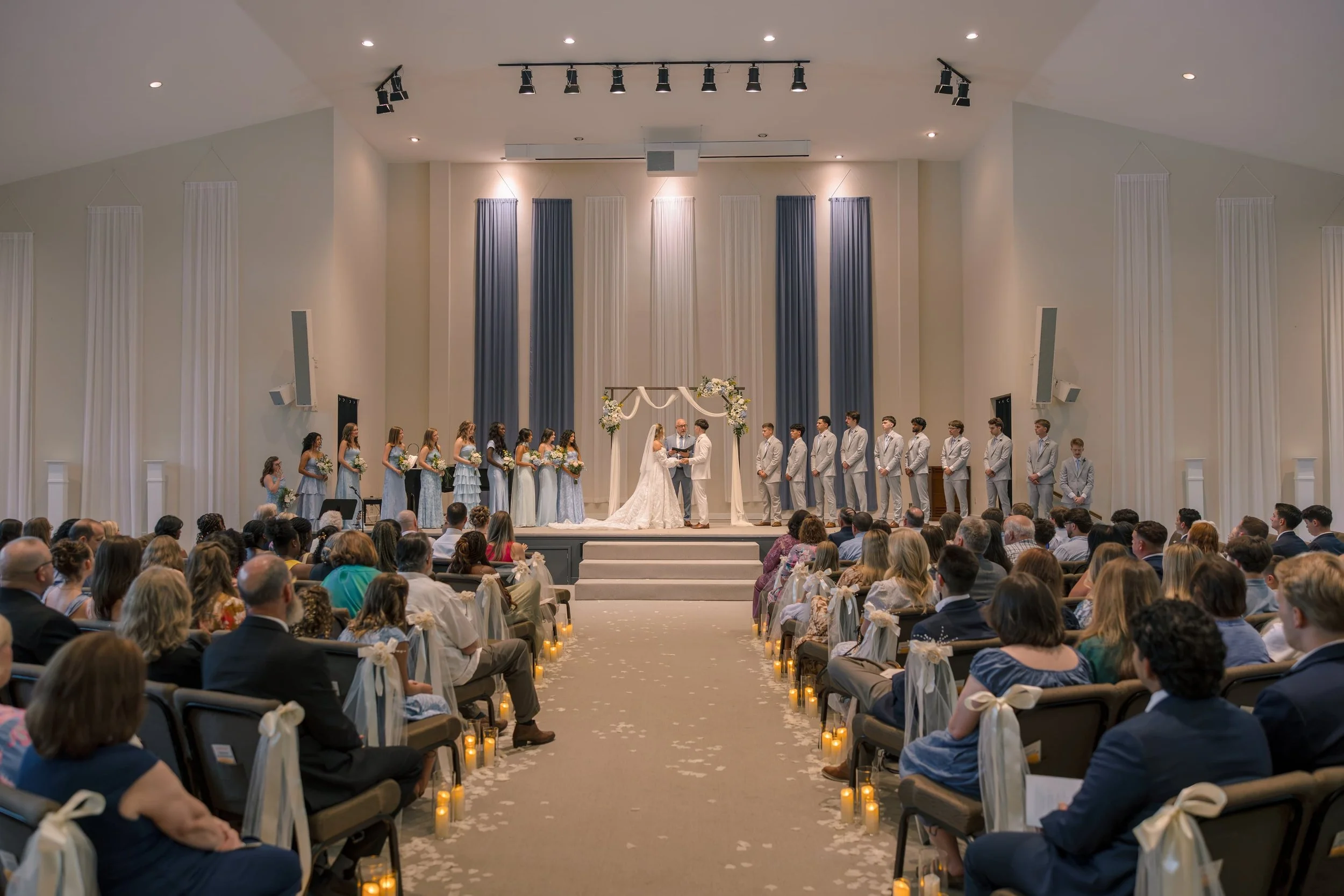 A wedding ceremony taking place inside a spacious, well-lit church or hall. The bride and groom are kneeling at an altar decorated with flowers and white fabric. Bridesmaids and groomsmen stand in a line on either side. The audience is seated in chai