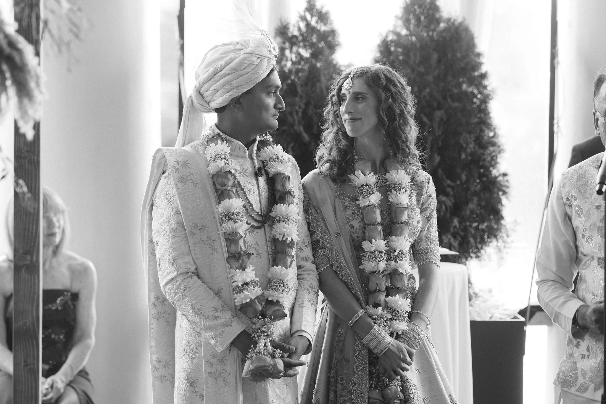 A black and white photo of a wedding ceremony, featuring a bride and groom dressed in traditional Indian attire, standing and holding hands, wearing floral garlands, with an outdoor scene of trees in the background.