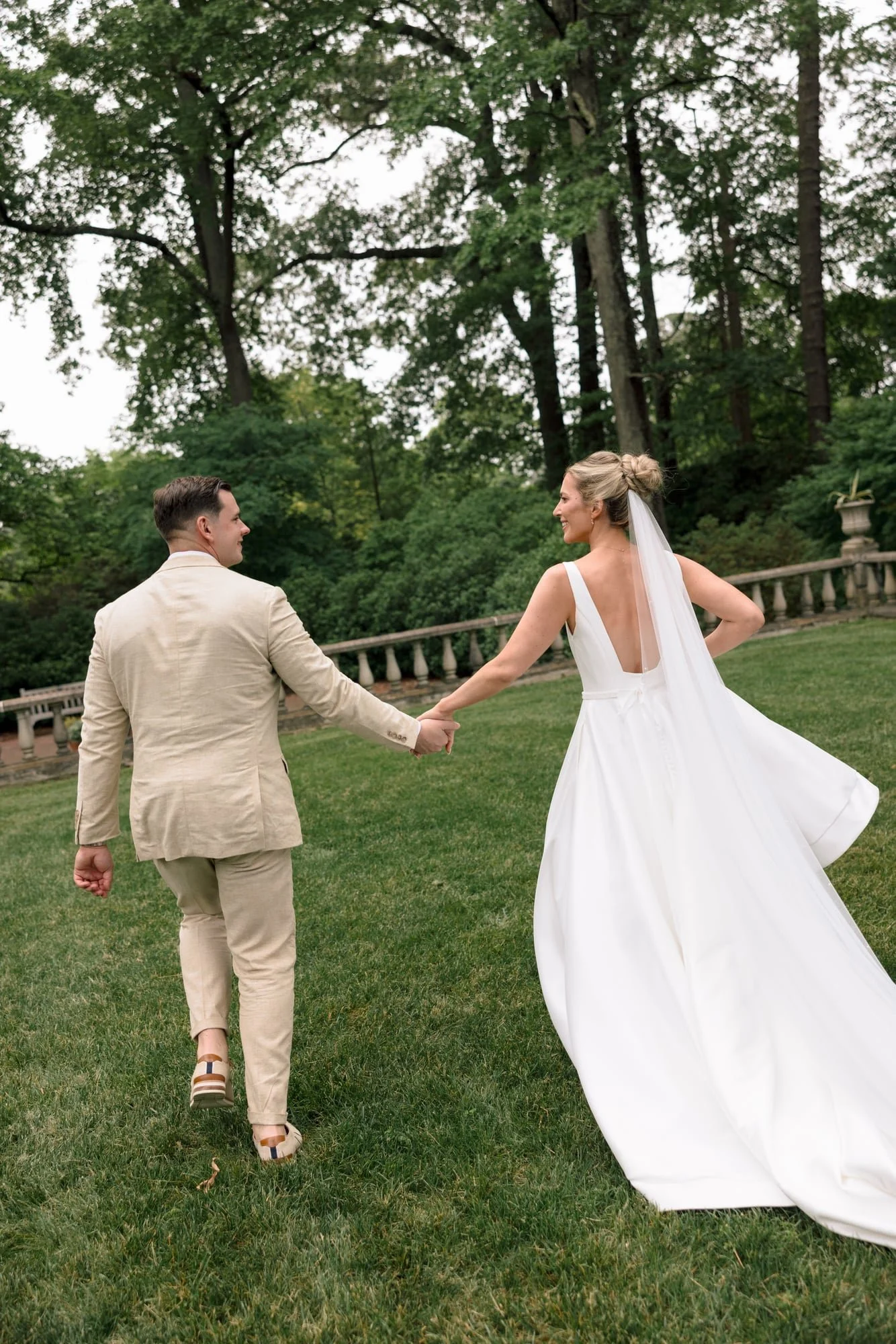 A couple in wedding attire holding hands and walking on a grassy lawn with lush green trees and a stone fence in the background.