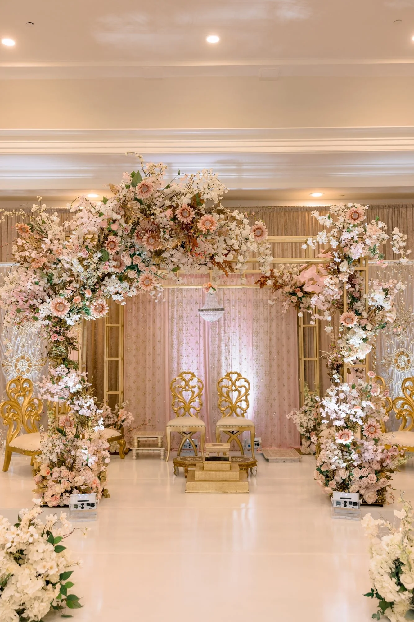 Elegant wedding ceremony setup with gold chairs, pink and white floral arrangements, and a chandelier in the background.