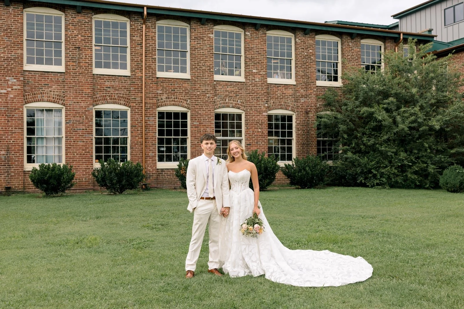 A bride and groom standing on a green lawn in front of a brick building. The bride is wearing a strapless white wedding gown and holding a bouquet, while the groom is dressed in a beige suit with a tie. They are holding hands and smiling.