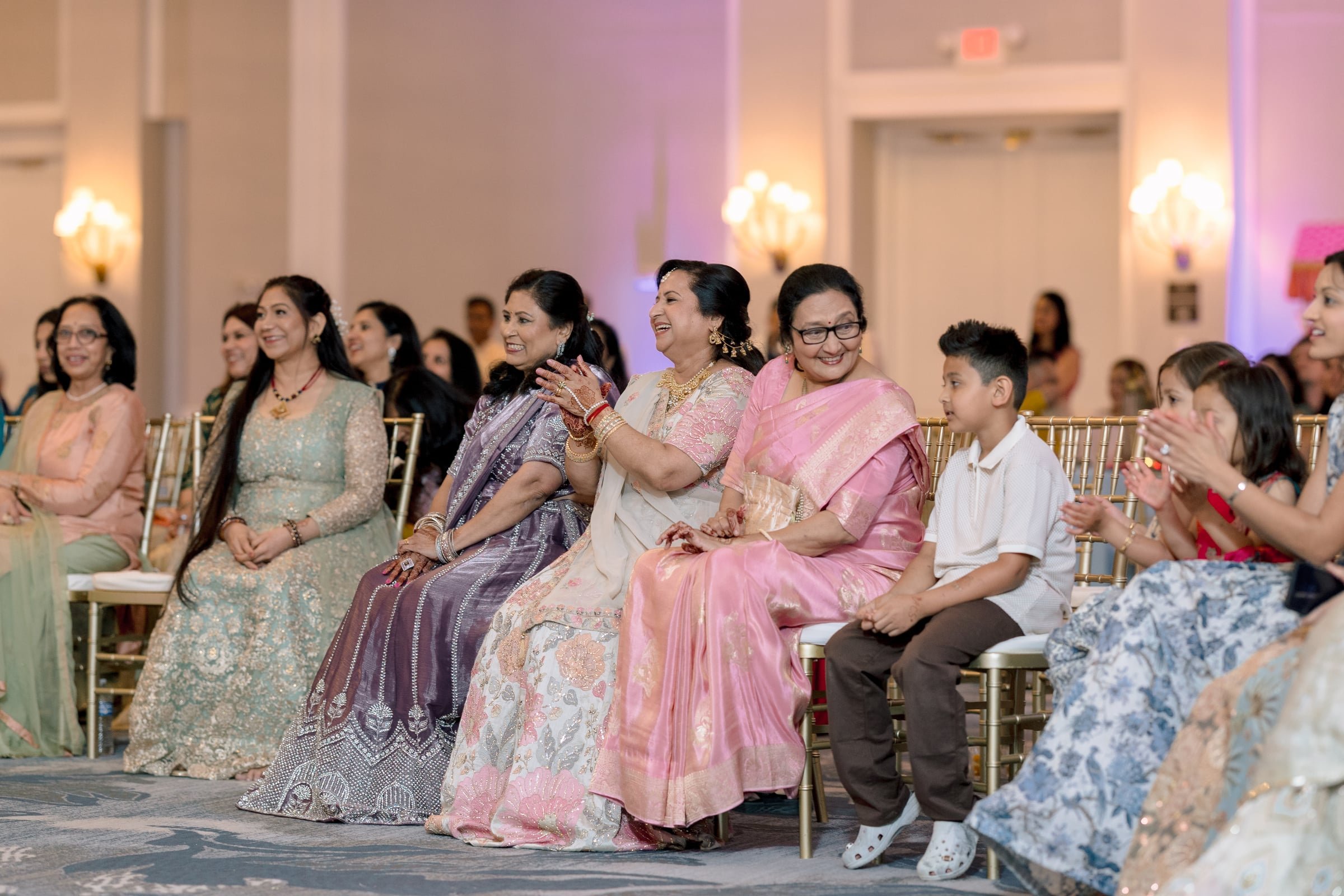 People sitting on chairs at an indoor event, mostly women wearing colorful traditional Indian dresses, some are smiling and clapping.