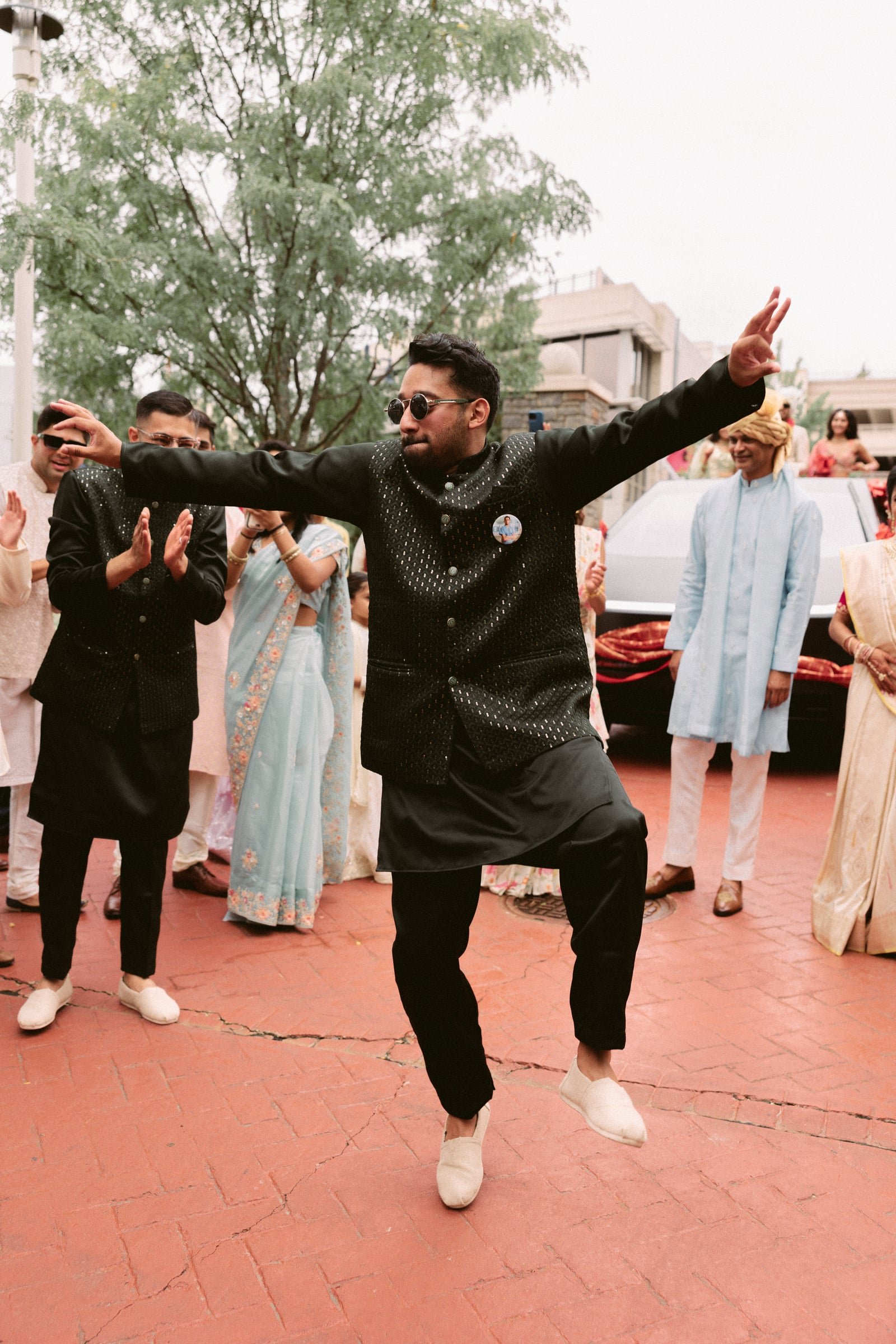 A man in black traditional Indian attire with sunglasses dancing at an outdoor celebration, surrounded by people in traditional Indian clothing.