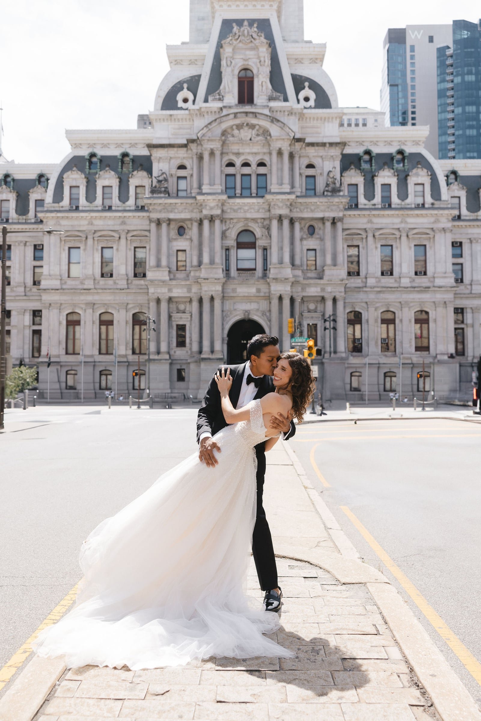 A couple in wedding attire embracing and smiling on a city street, with a historic building and modern skyscrapers in the background.