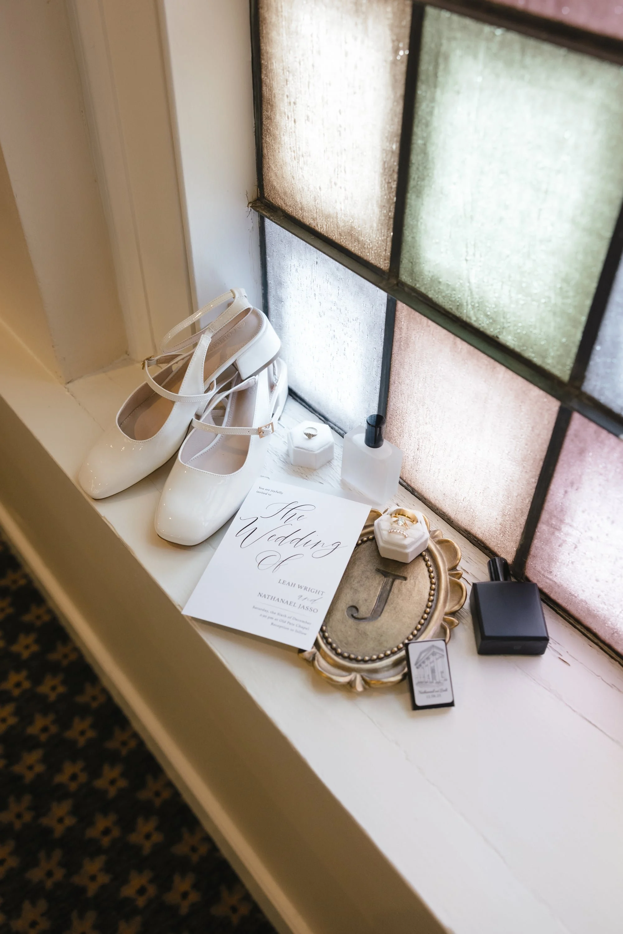 A white pair of wedding shoes, a wedding invitation, and decorative items placed on a windowsill next to a stained glass window.