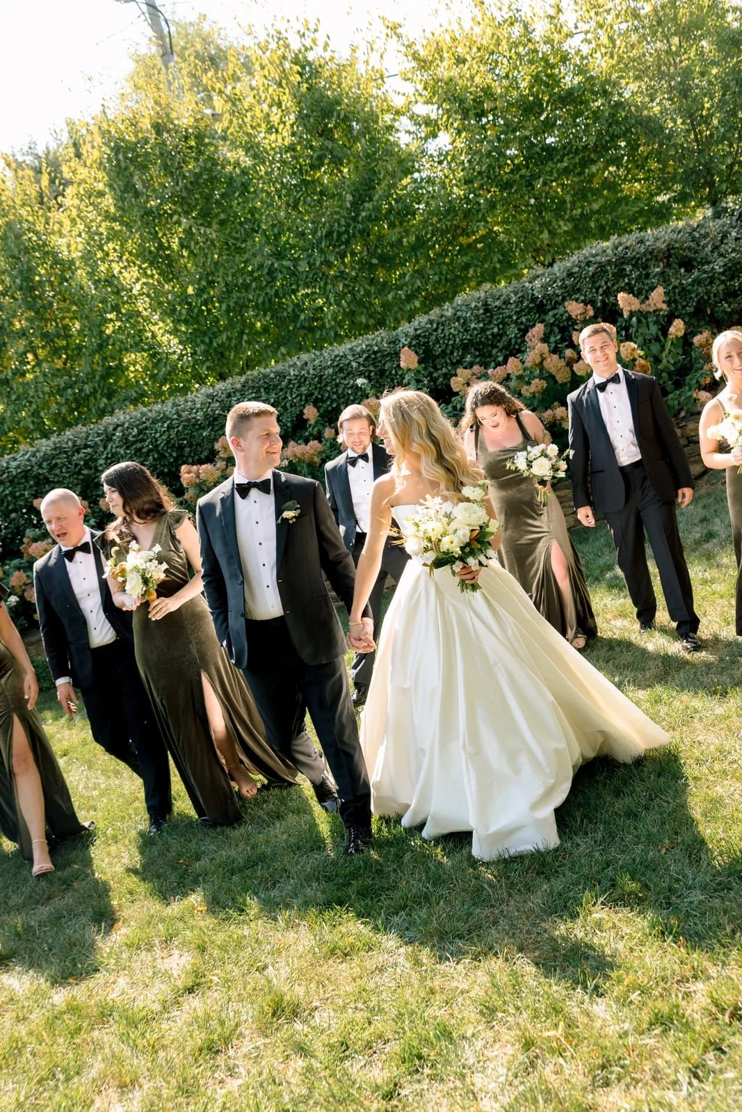 A wedding party walking outdoors on grass, with a bride in a white dress and veil holding a bouquet, and a groom in a black tuxedo, surrounded by bridesmaids and groomsmen in formal attire, smiling and holding flowers.