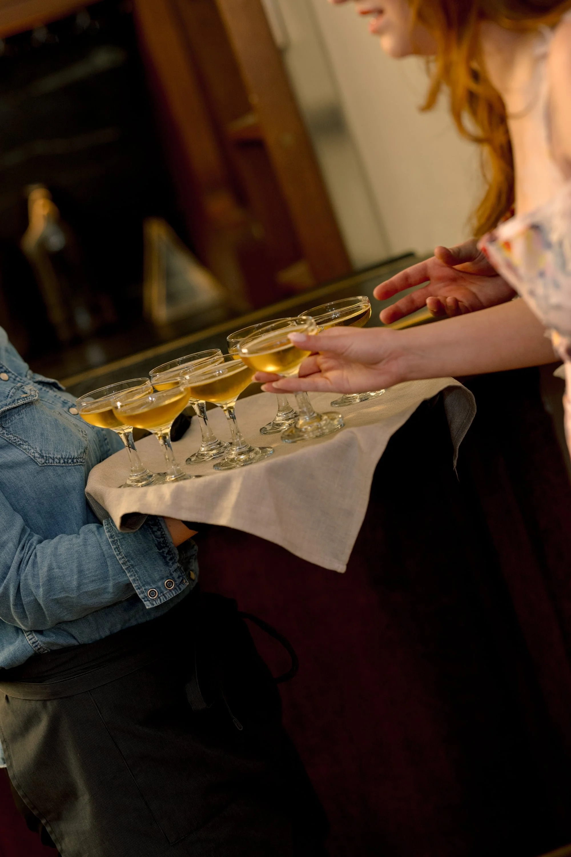 Person wearing a denim jacket and gray pants serving multiple glasses of a yellow beverage, possibly champagne or wine, on a beige cloth.