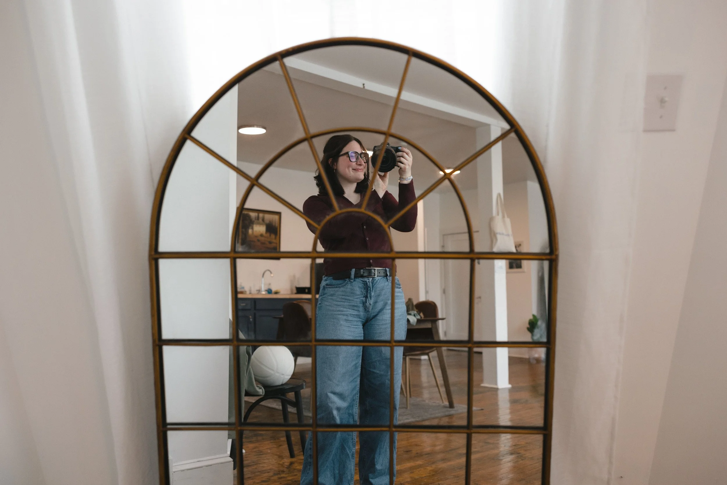 A woman with glasses takes a selfie in a decorative mirror, standing in a room with wooden floors and white walls.