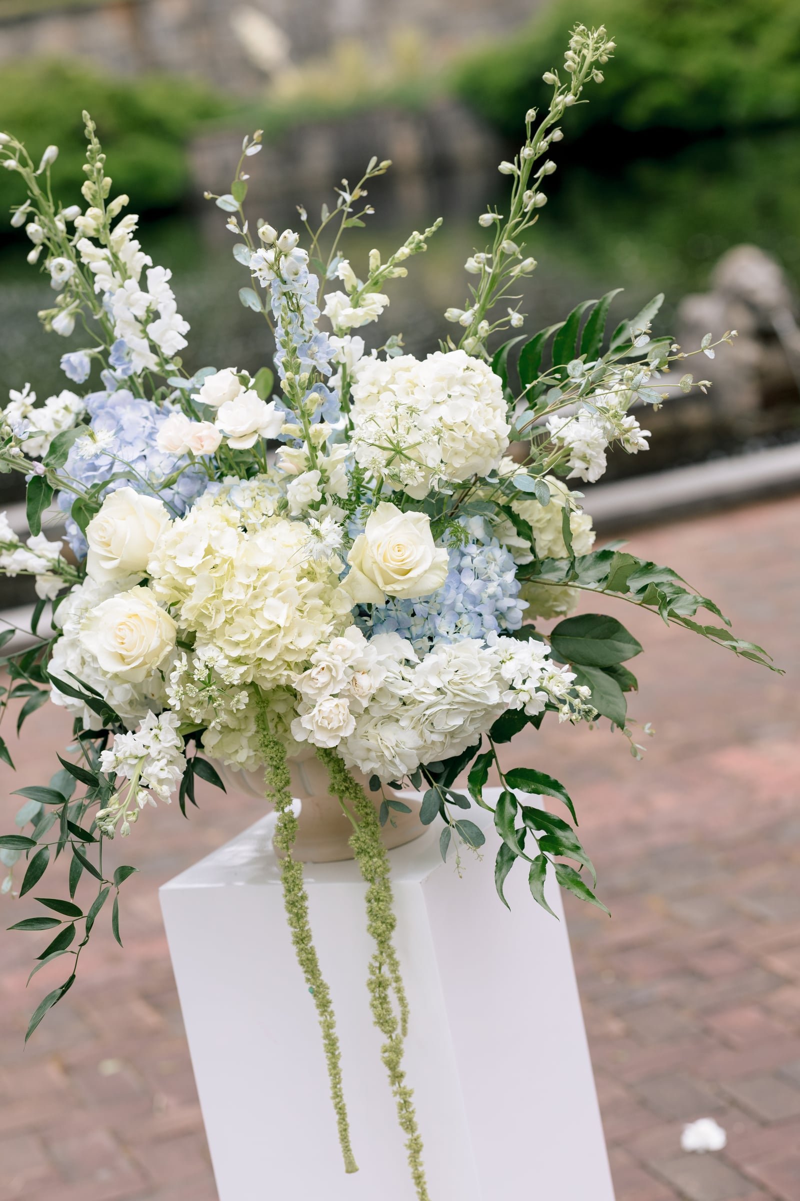 An elegant floral arrangement with white roses, hydrangeas, and greenery, placed on a white pedestal outdoors.
