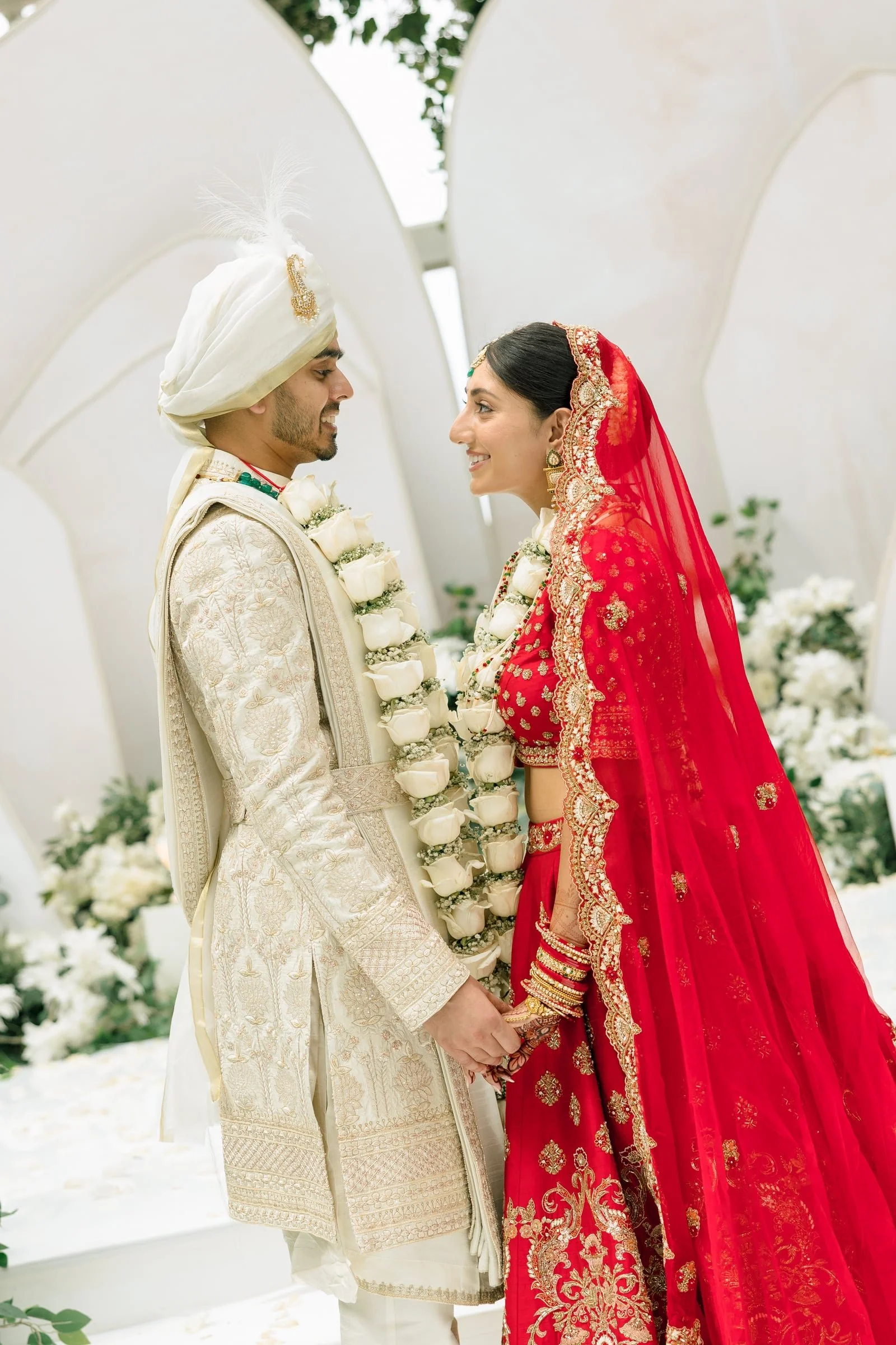 A bride and groom in traditional Indian wedding attire holding hands during a wedding ceremony, smiling at each other, with floral decorations in the background.