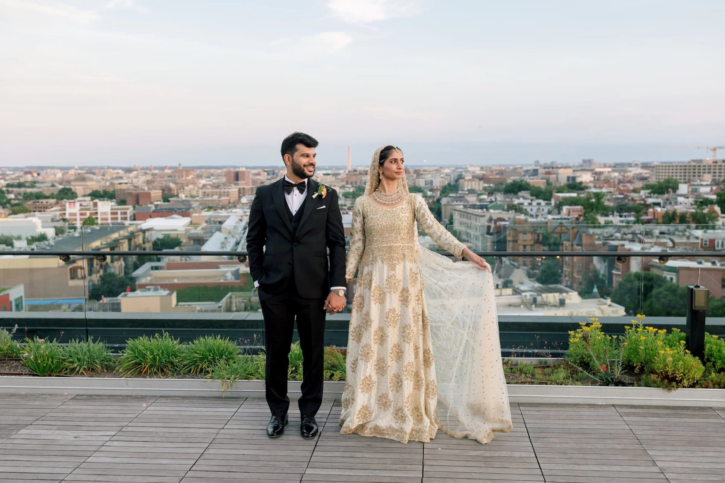 A bride and groom holding hands on a rooftop with a city skyline background, with the groom dressed in a black tuxedo and the bride dressed in a gold and cream traditional gown.