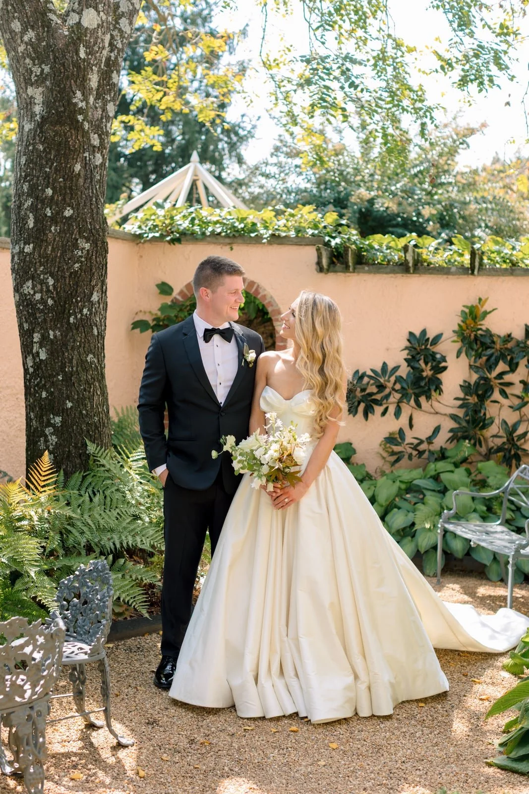 A bride and groom standing together outdoors on a wedding day, smiling at each other. The bride is holding a bouquet of white flowers, and the groom is wearing a black tuxedo with a bow tie. They are surrounded by greenery and a beige wall with plants.