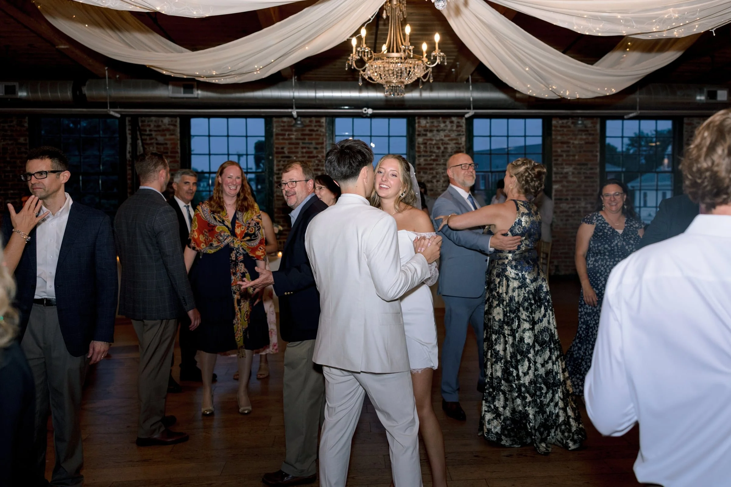 People dancing and socializing at a wedding reception or celebration in a decorated indoor venue with a chandelier and draped fabric ceiling.