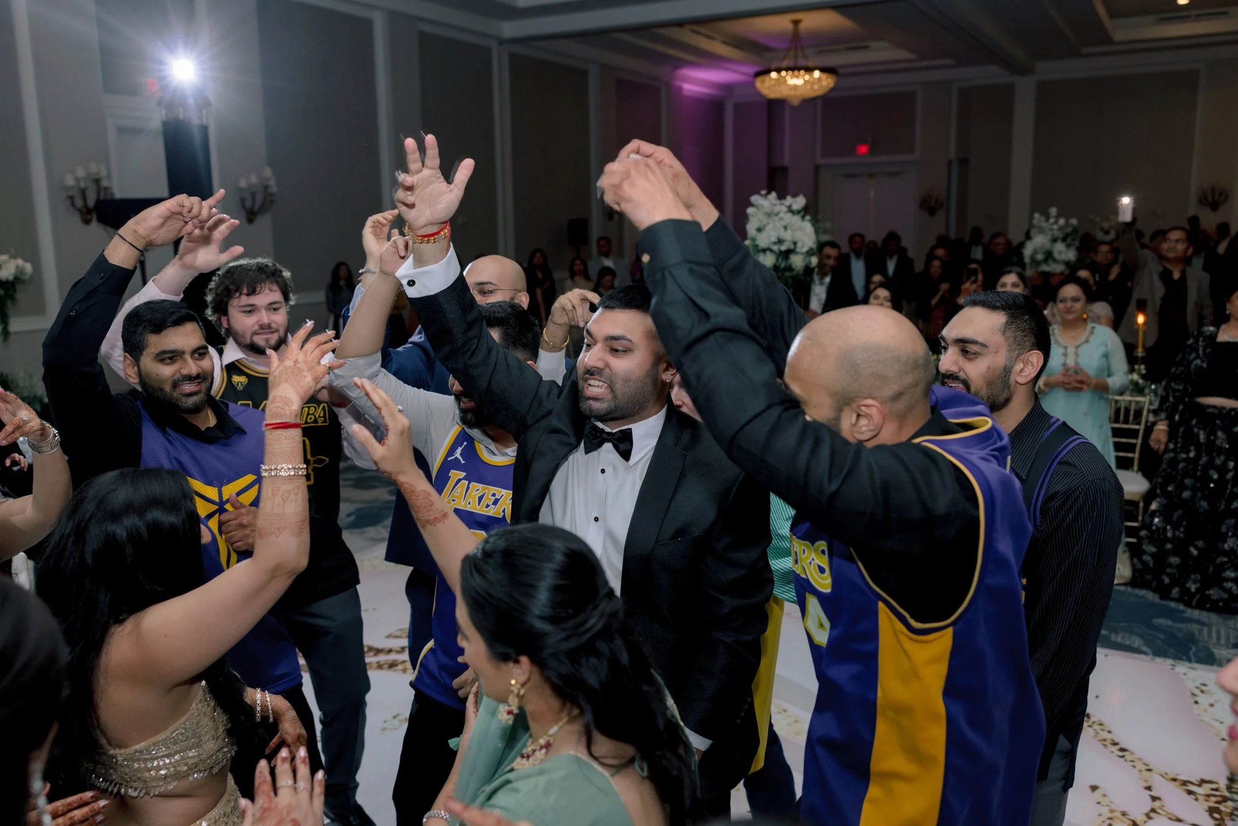 A group of people dancing and celebrating at a wedding reception in a decorated banquet hall. Some are wearing sports jerseys, others are dressed in formal attire.