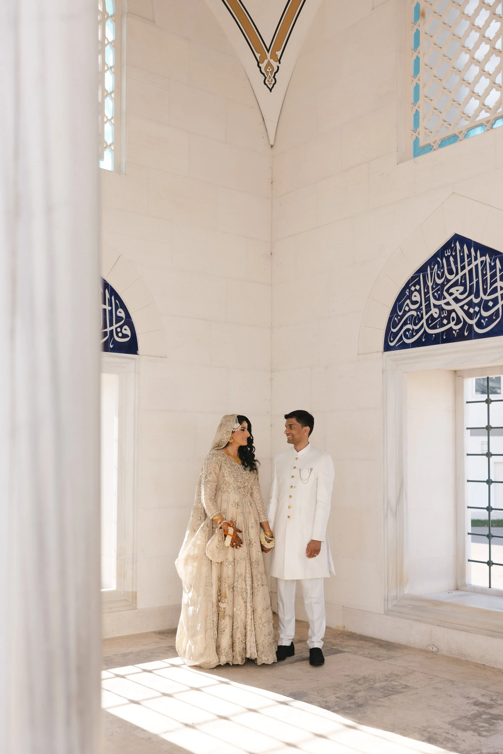 A couple dressed in traditional South Asian wedding attire standing in a bright, white room with high ceilings, ornate architectural details, and Islamic calligraphy. The woman wears a gold and beige embroidered lehenga with jewelry and a veil, and t