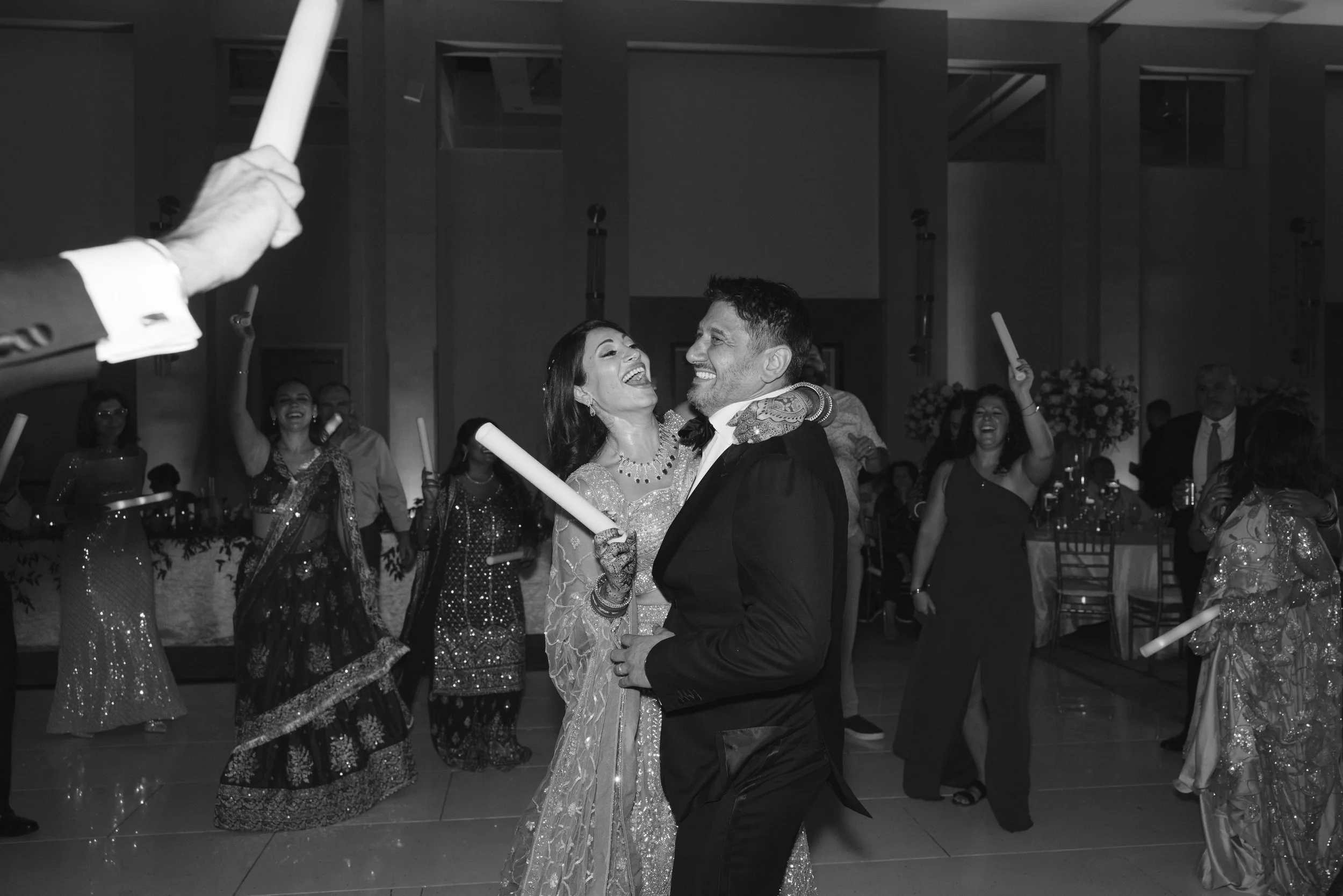 A wedding celebration with a bride and groom dancing, surrounded by guests in formal attire, in an indoor venue with decorative lighting and flower arrangements.