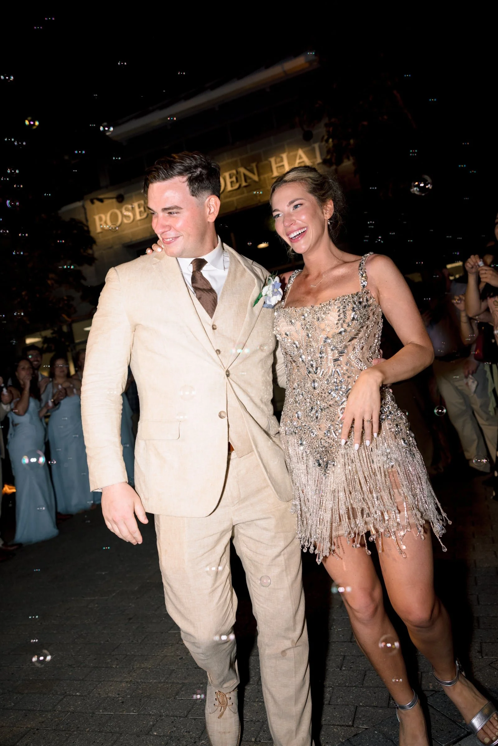A couple dressed in formal attire celebrating at a wedding reception. The man wears a beige suit with a white shirt and brown tie. The woman wears a sparkly, embellished cocktail dress. They are smiling and standing close together, surrounded by gues