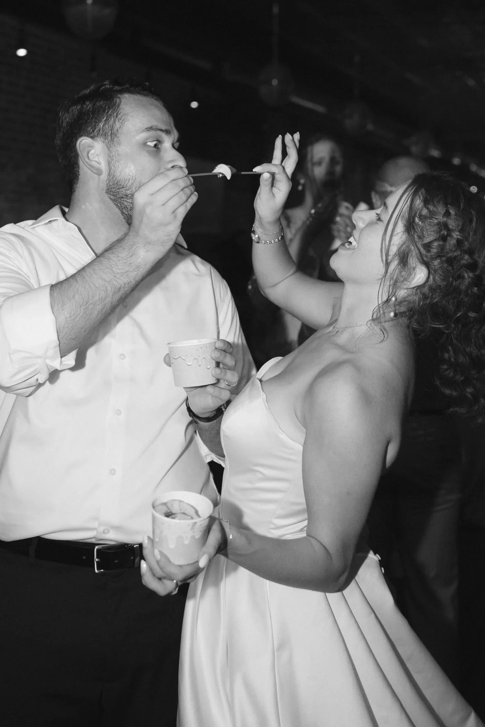 A black and white photo of a man and woman at a celebration, with the man holding a cup and the woman holding a skewer with a marshmallow while smiling at each other.