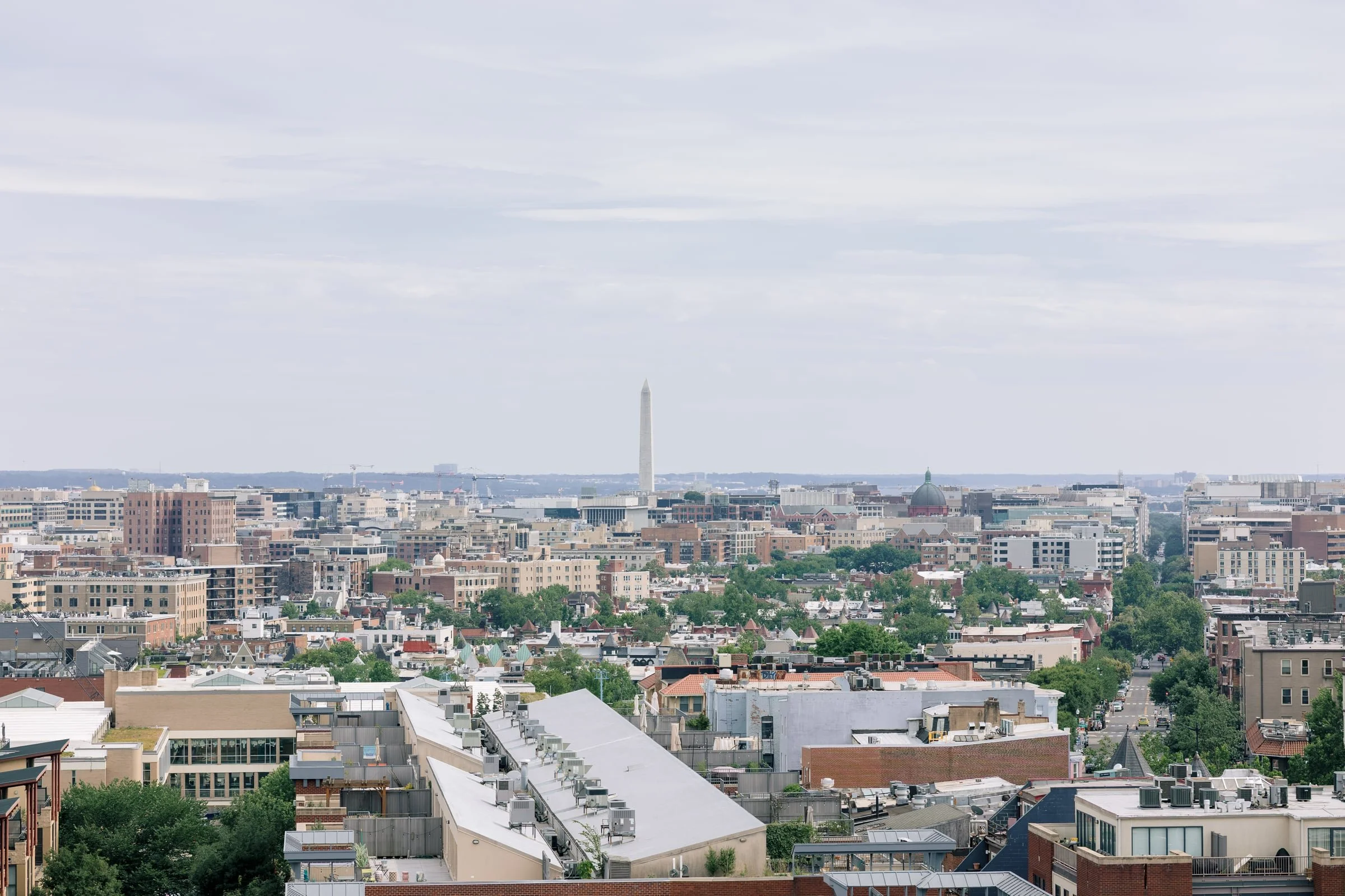 Cityscape view of Washington, D.C., with the Washington Monument visible in the distance under a cloudy sky.