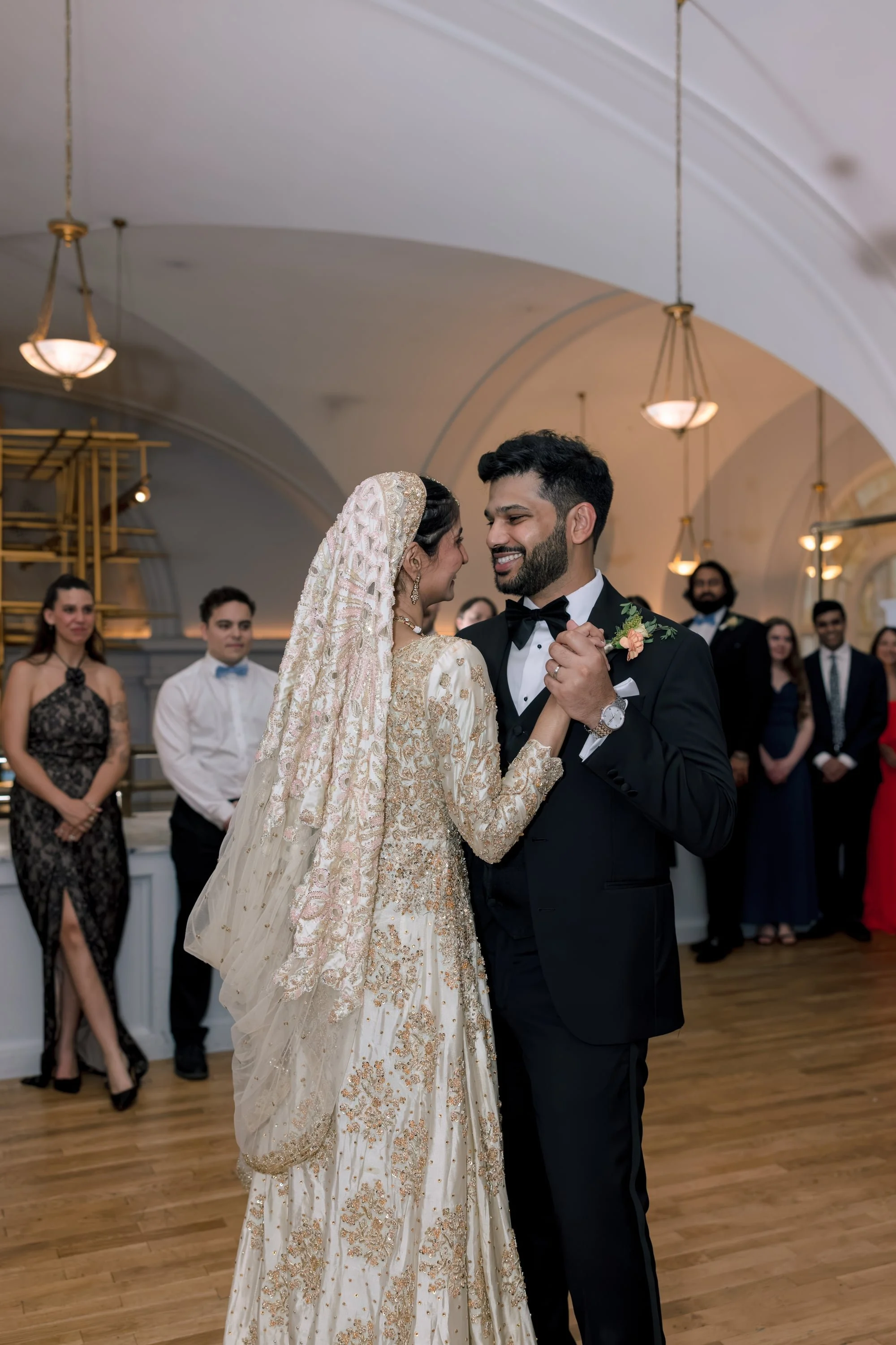 A bride and groom dance together at her wedding reception, with wedding guests watching in the background inside a decorated venue.