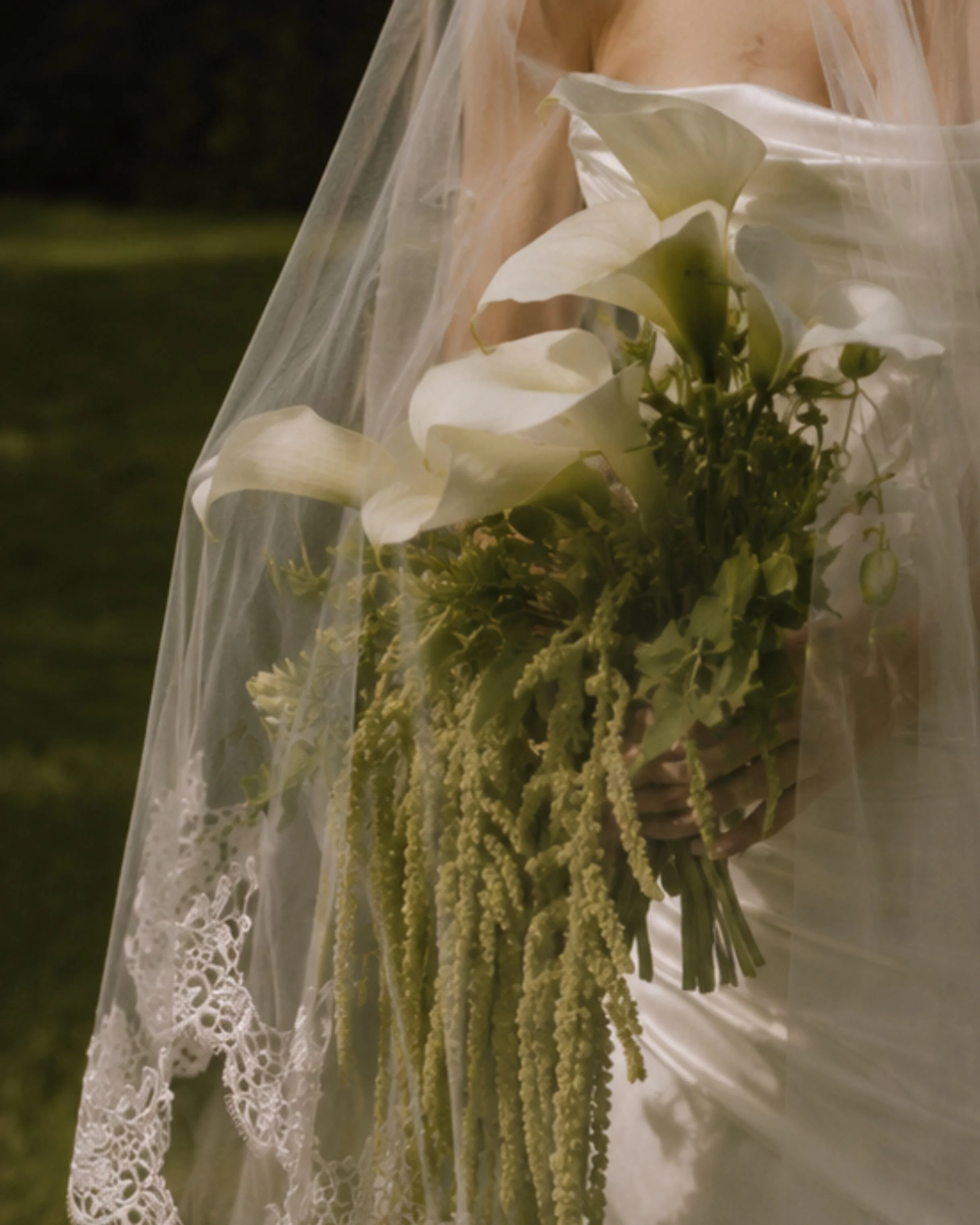 A bouquet of white calla lilies wrapped in lace and clear plastic, held against a dark, out-of-focus background.