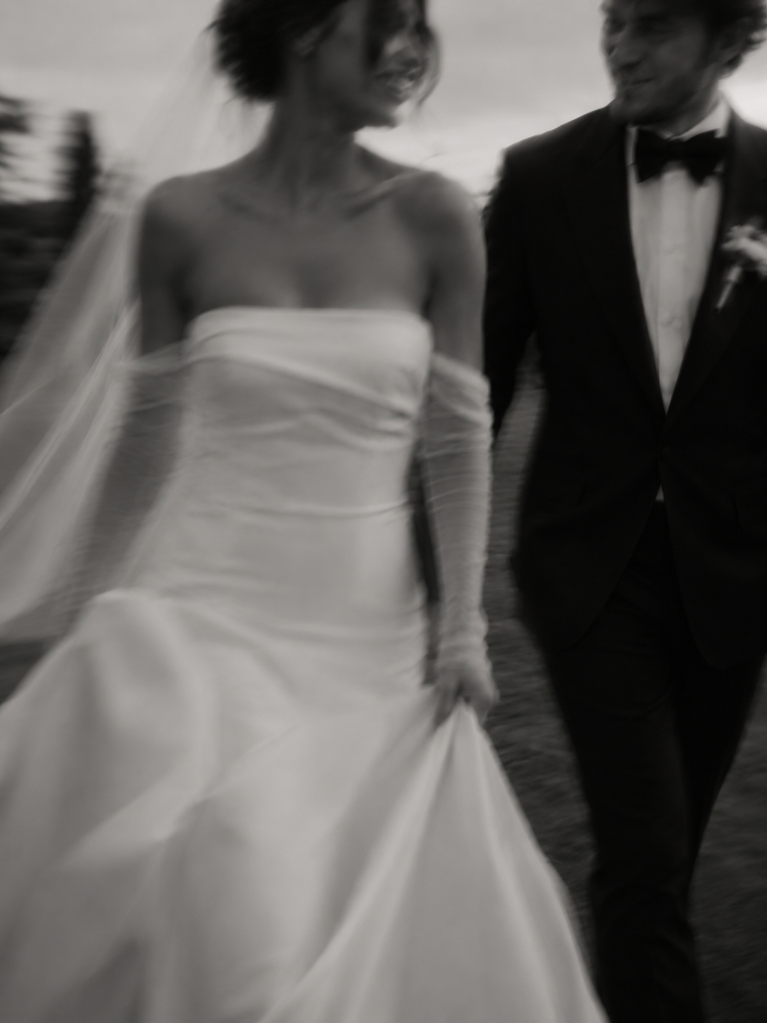 A bride and groom walking together on their wedding day, with the bride wearing a strapless wedding gown and the groom dressed in a tuxedo, holding hands and smiling.
