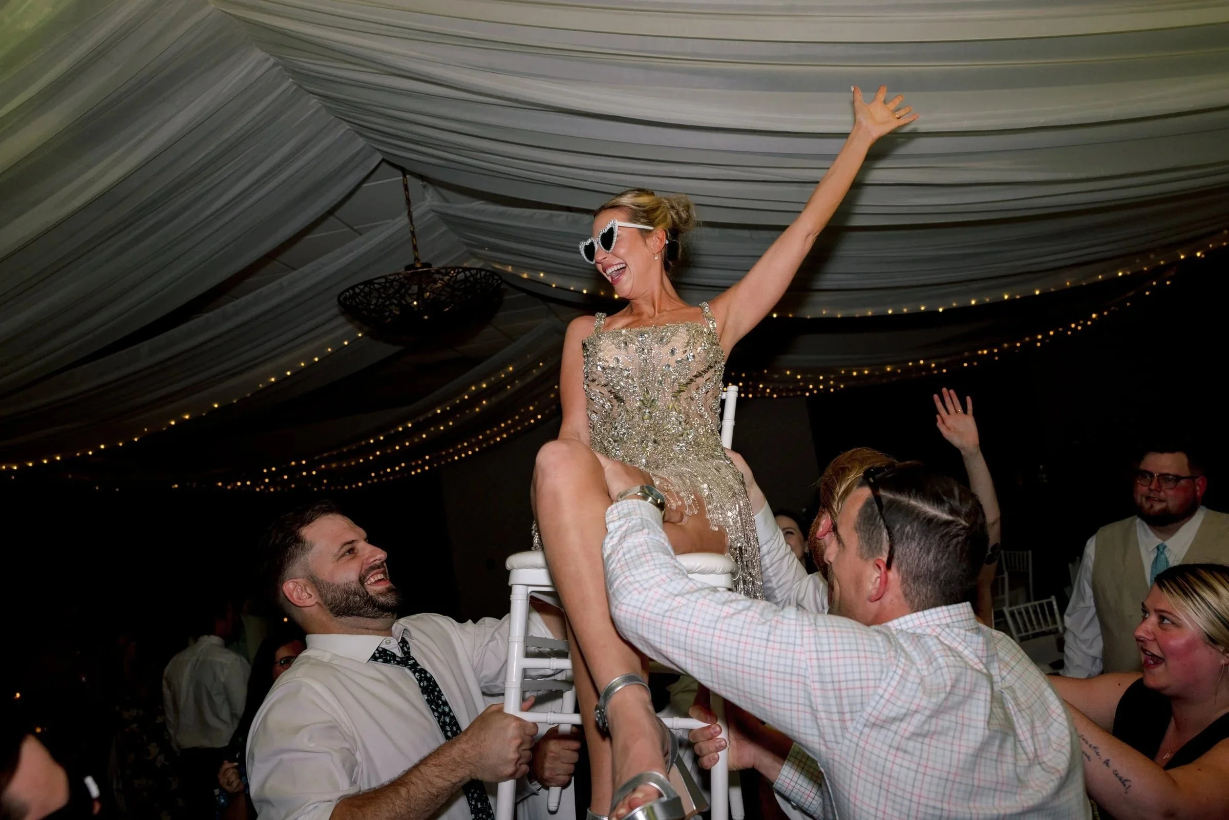 A woman wearing sunglasses and a glittery dress is sitting on top of people who are standing in a celebration or party, with a decorated ceiling and string lights in the background.