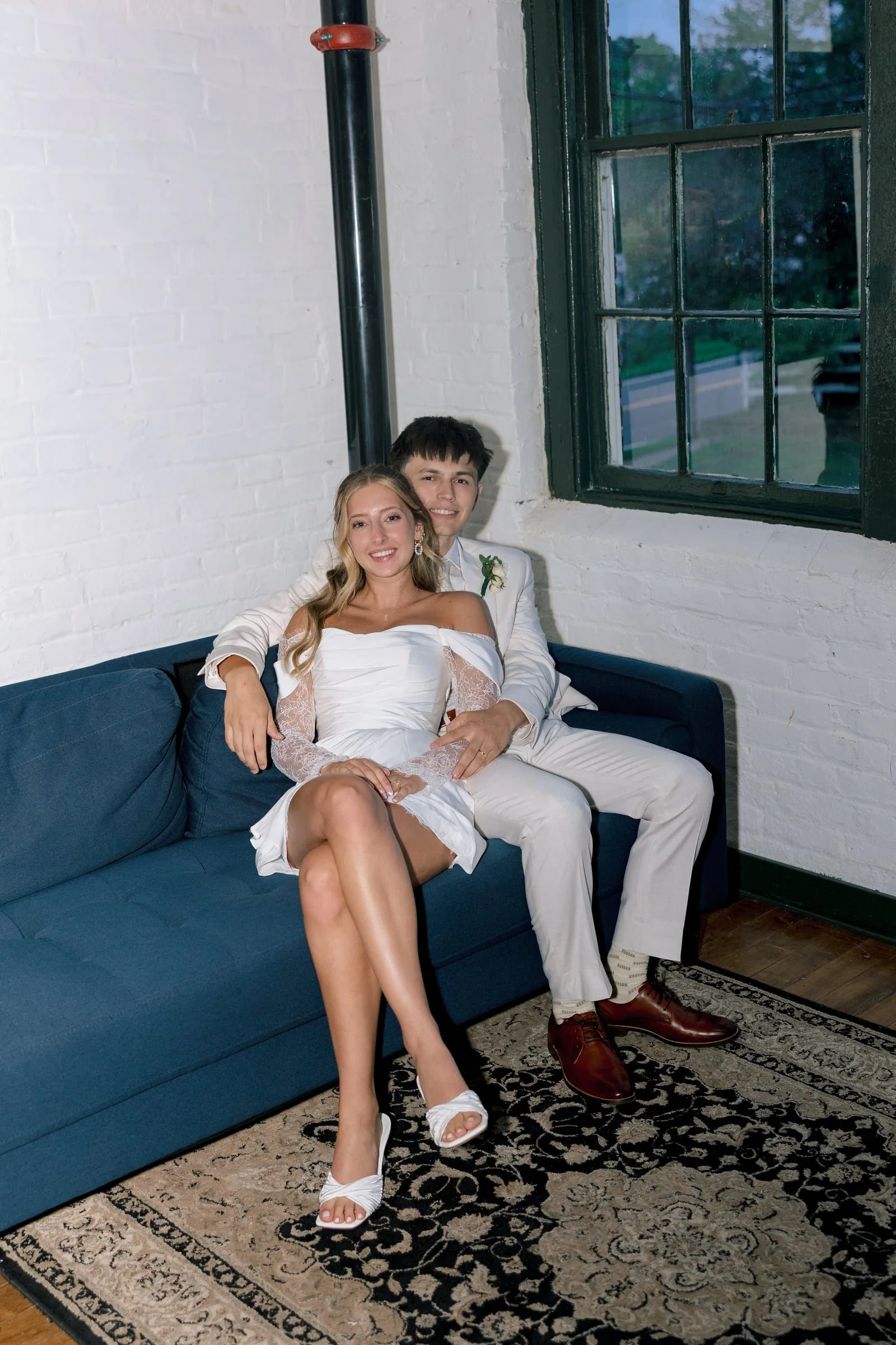 A young couple dressed in wedding attire sitting on a blue sofa in a room with white brick walls, a large window, and a patterned rug, smiling and posing for the photo.