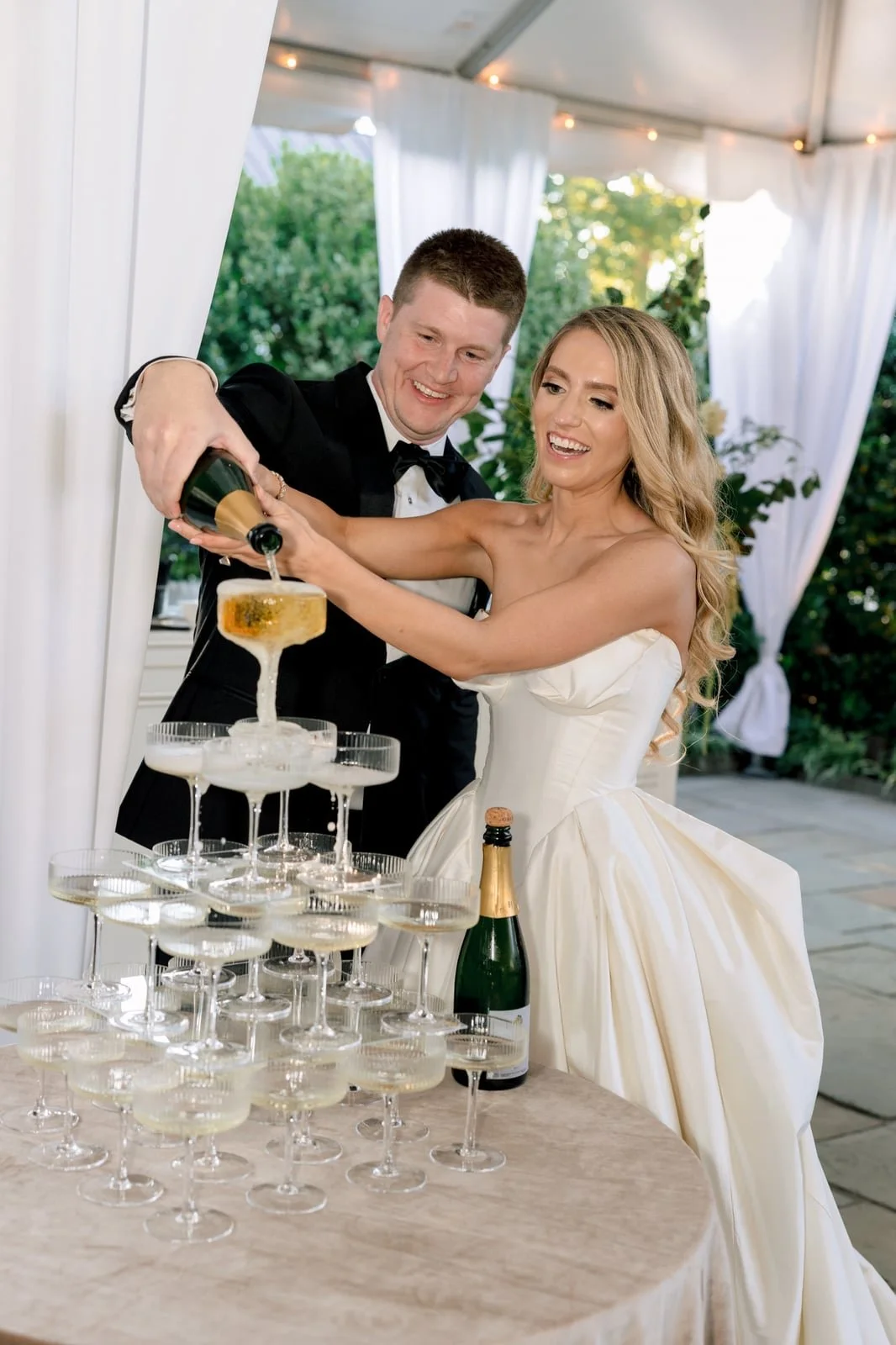 A bride and groom pour champagne into a tower of glasses at their wedding reception outdoors, with lush green trees and white drapery in the background.