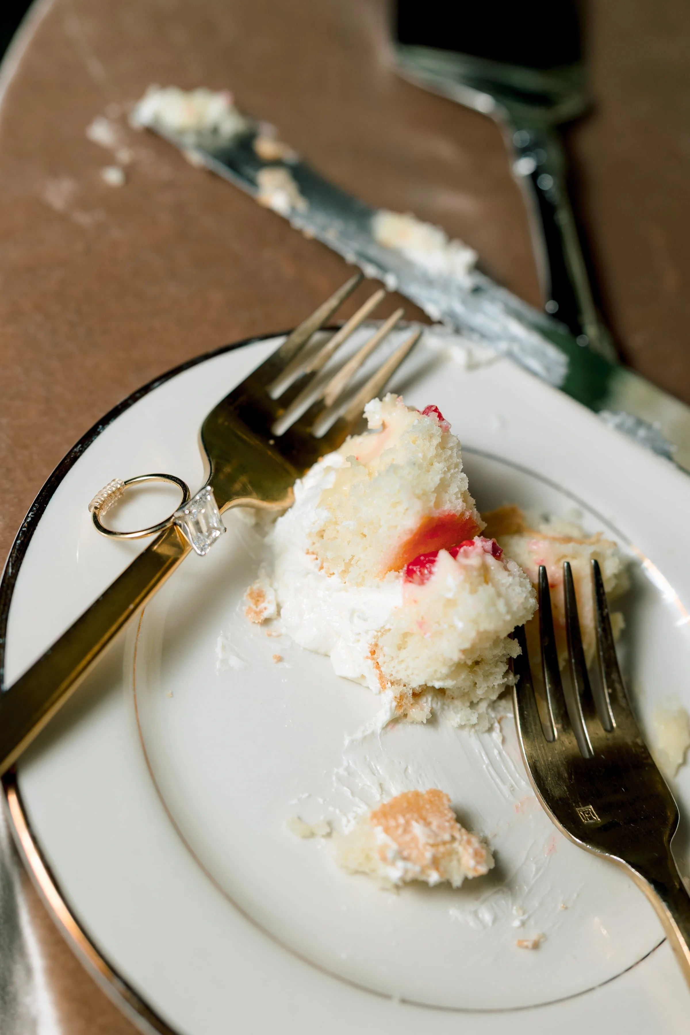 A white plate with a piece of partially eaten white cake with red fruit topping, a gold fork with decorative handle, and a gold butter knife, with a fork and knife placed on the plate.