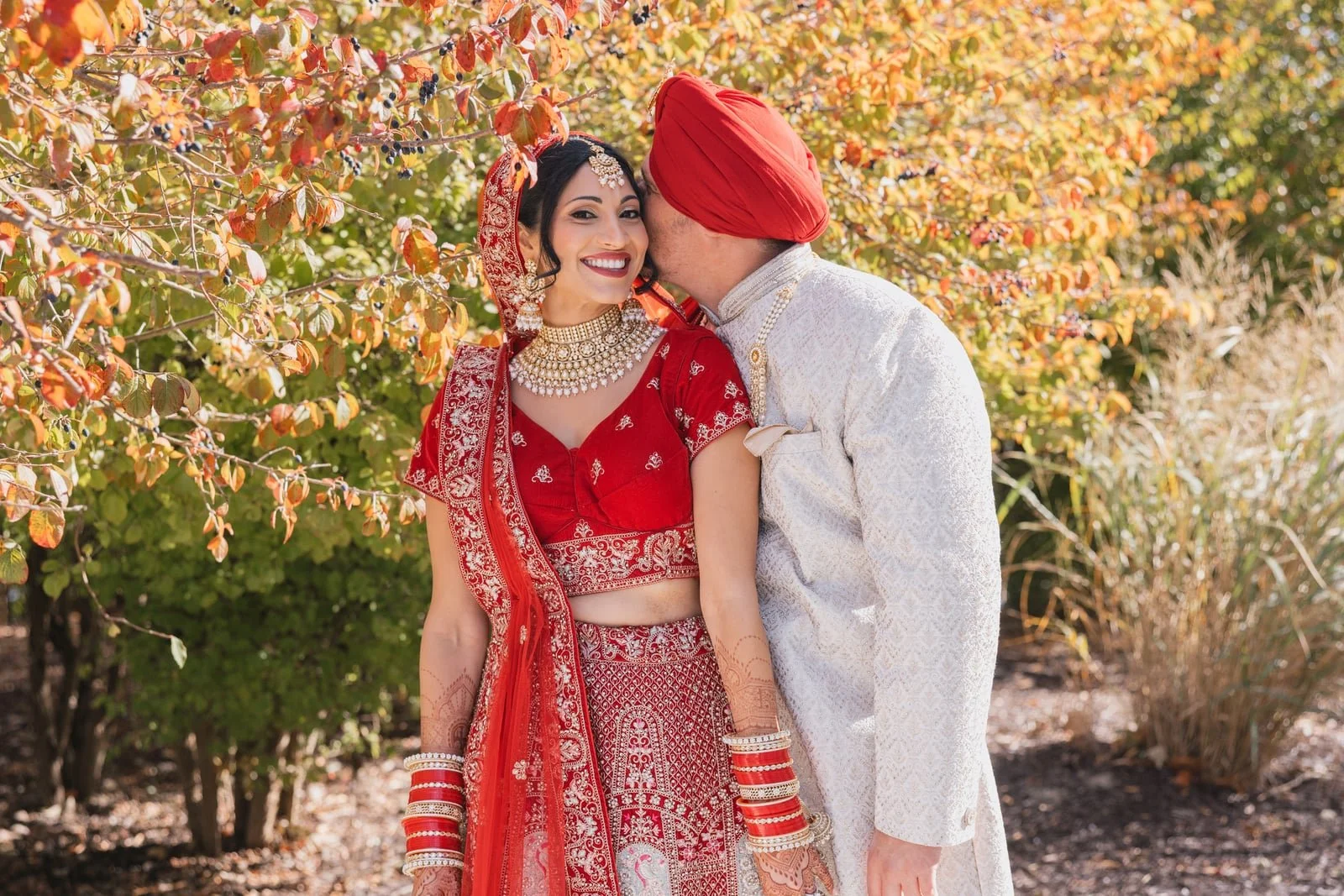 A couple dressed in traditional Indian wedding attire standing outdoors, with a woman in red and jewelry smiling and a man whispering to her, surrounded by autumn foliage.