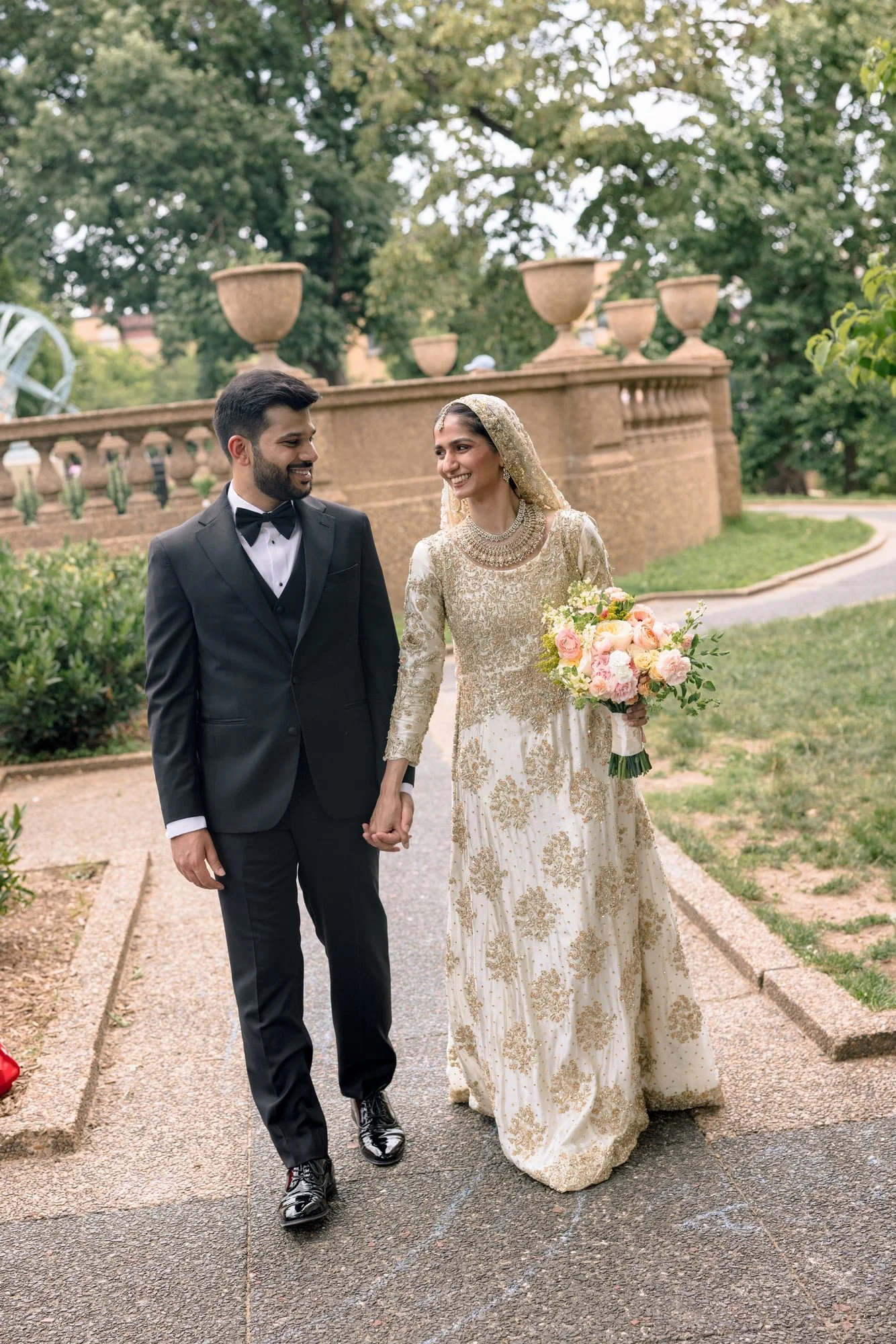 A bride and groom walking hand-in-hand outdoors, the bride wearing a white and gold wedding dress with a headscarf and holding a bouquet, and the groom wearing a black tuxedo and bow tie, smiling at each other.