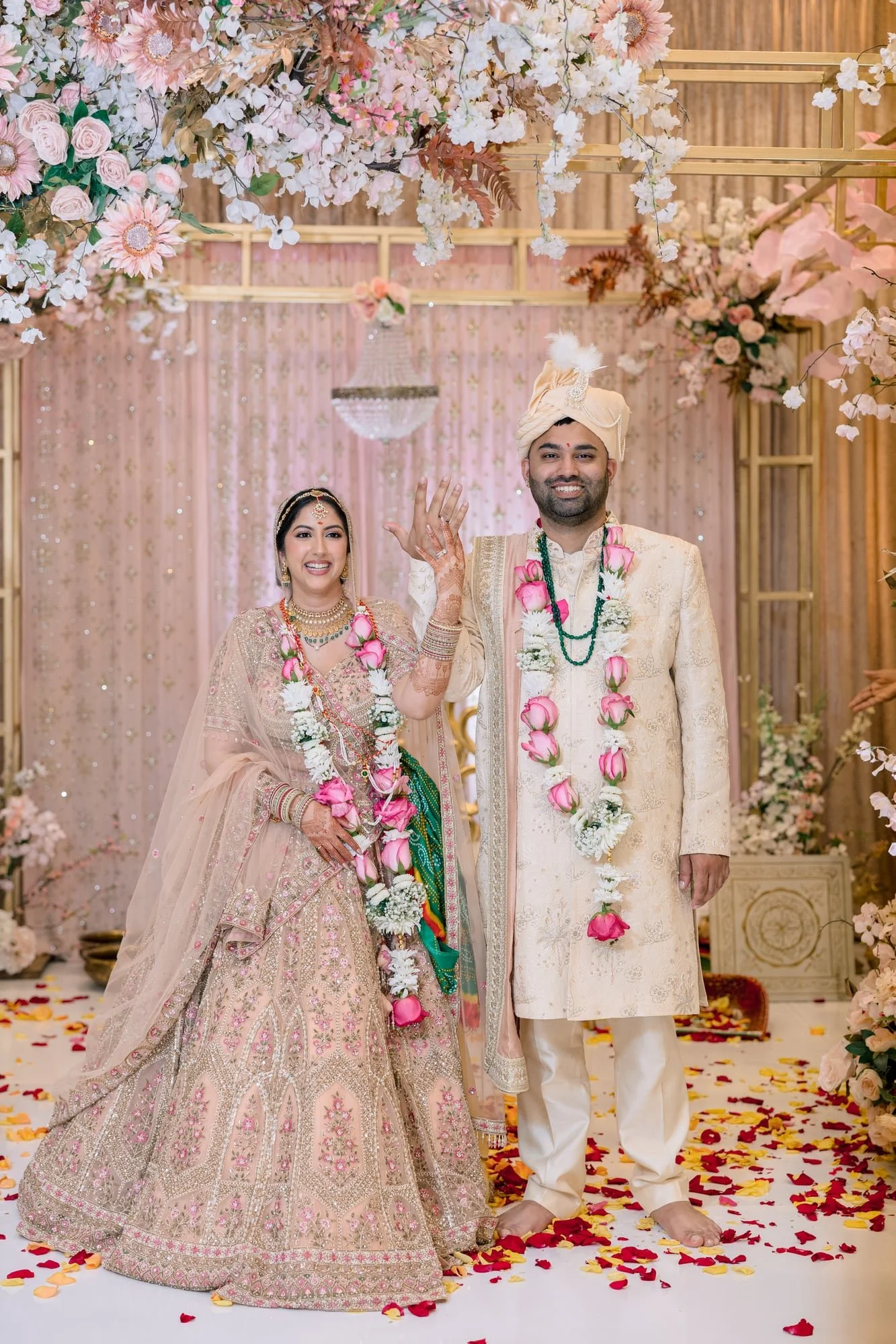 An Indian wedding ceremony with a bride and groom standing underneath a floral canopy. The bride wears an ornate pink and gold bridal lehenga and jewelry, and the groom wears a cream-colored sherwani and turban. They are smiling, holding hands, and surrounded by flower petals on the floor.