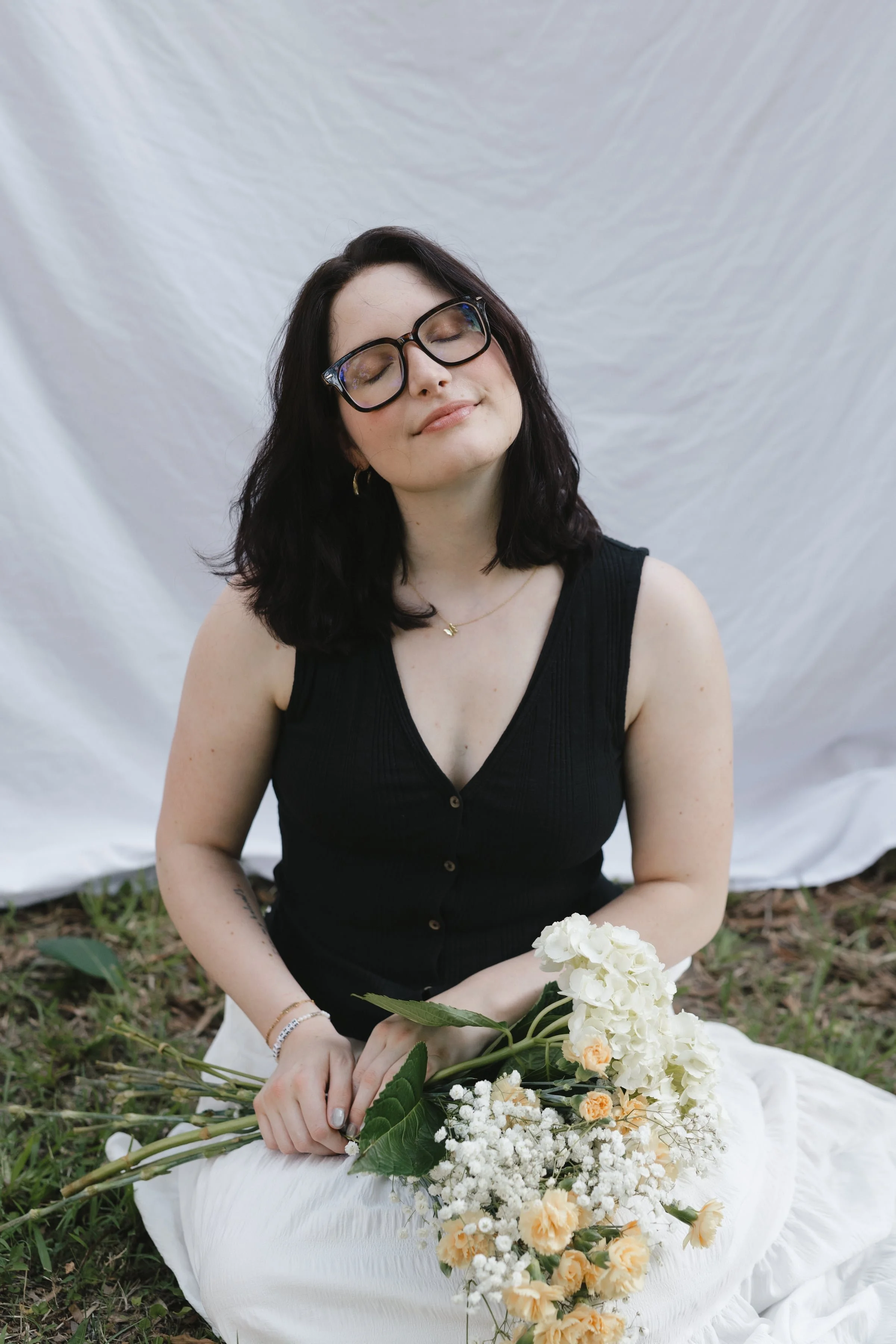 A young woman with dark hair and glasses, wearing a black sleeveless top, sitting outdoors on a white blanket with a bouquet of white and peach flowers, eyes closed, with a white backdrop behind her.