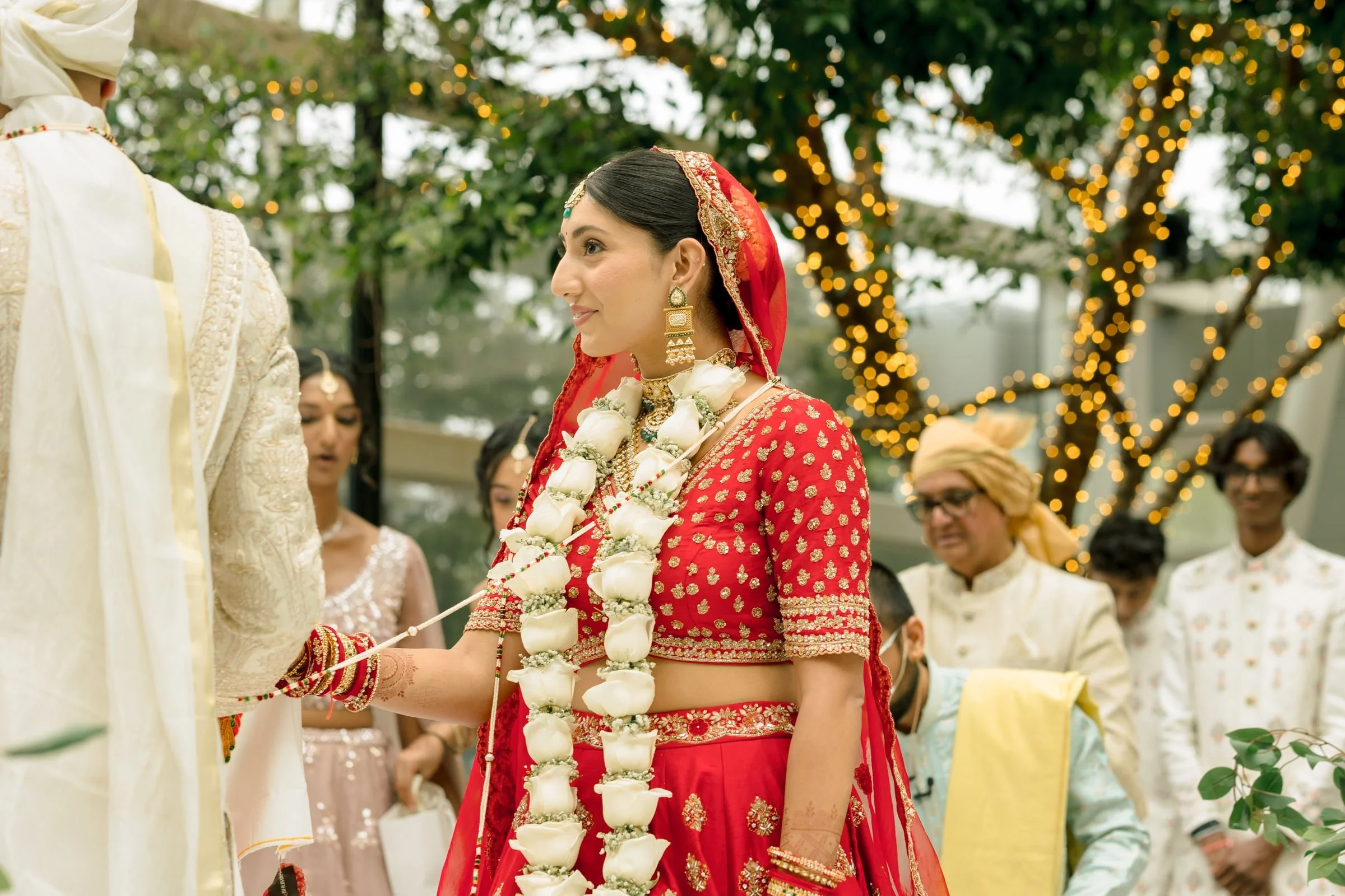 A woman wearing a red bridal outfit with gold embroidery and a floral necklace is participating in a traditional Indian wedding ceremony, holding a string. Other guests are visible in the background under lit trees.