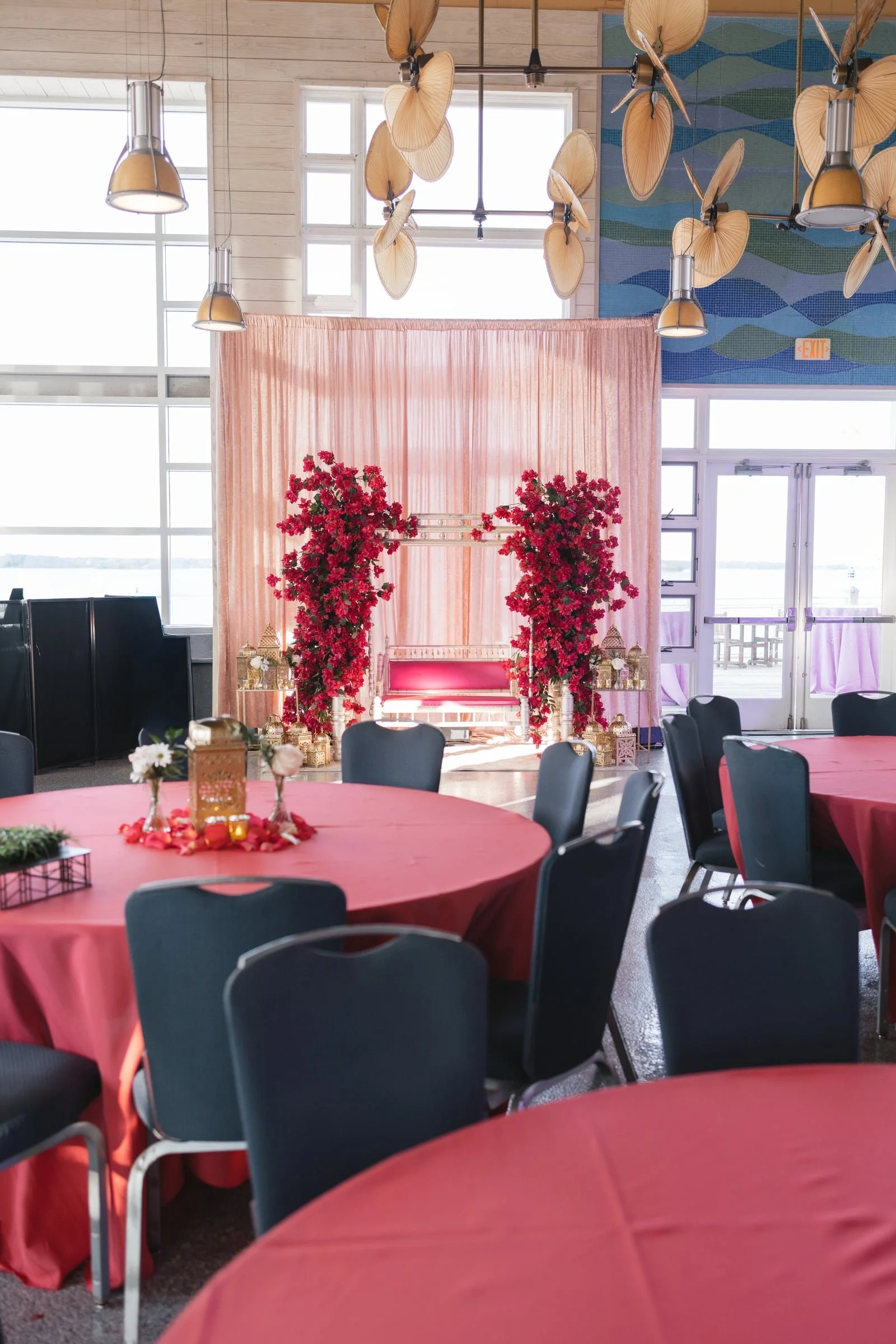 Indoor event space decorated with pink curtains, red floral arrangements, and round tables with pink tablecloths and blue chairs. There is a backdrop with pink flowers and gold lanterns, and large windows showing an outdoor view. Overhead, from the c