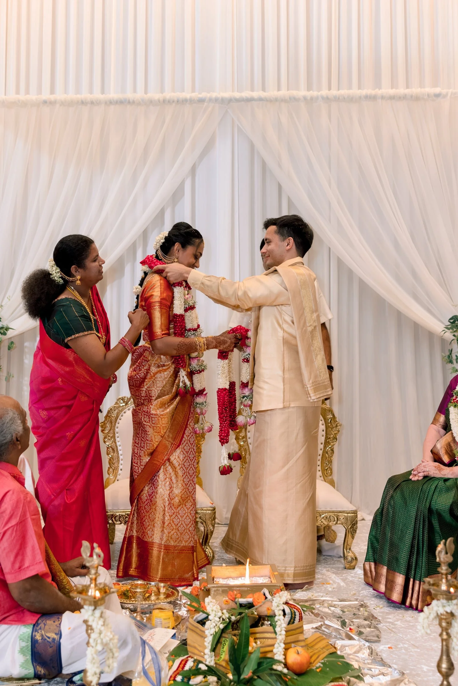 A traditional Indian wedding ceremony with the bride and groom dressed in ornate attire, participating in a ritual guided by two women, in a decorated indoor setting with white drapes and ceremonial items on the floor.