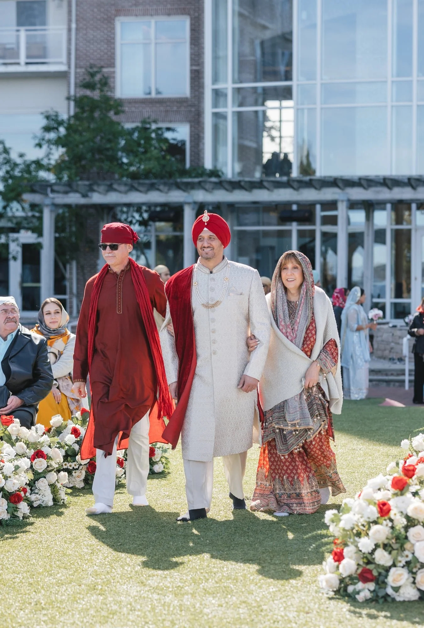 A diverse group of people, including Indian and Western individuals, walking together at a cultural wedding ceremony outdoors, with floral decorations.