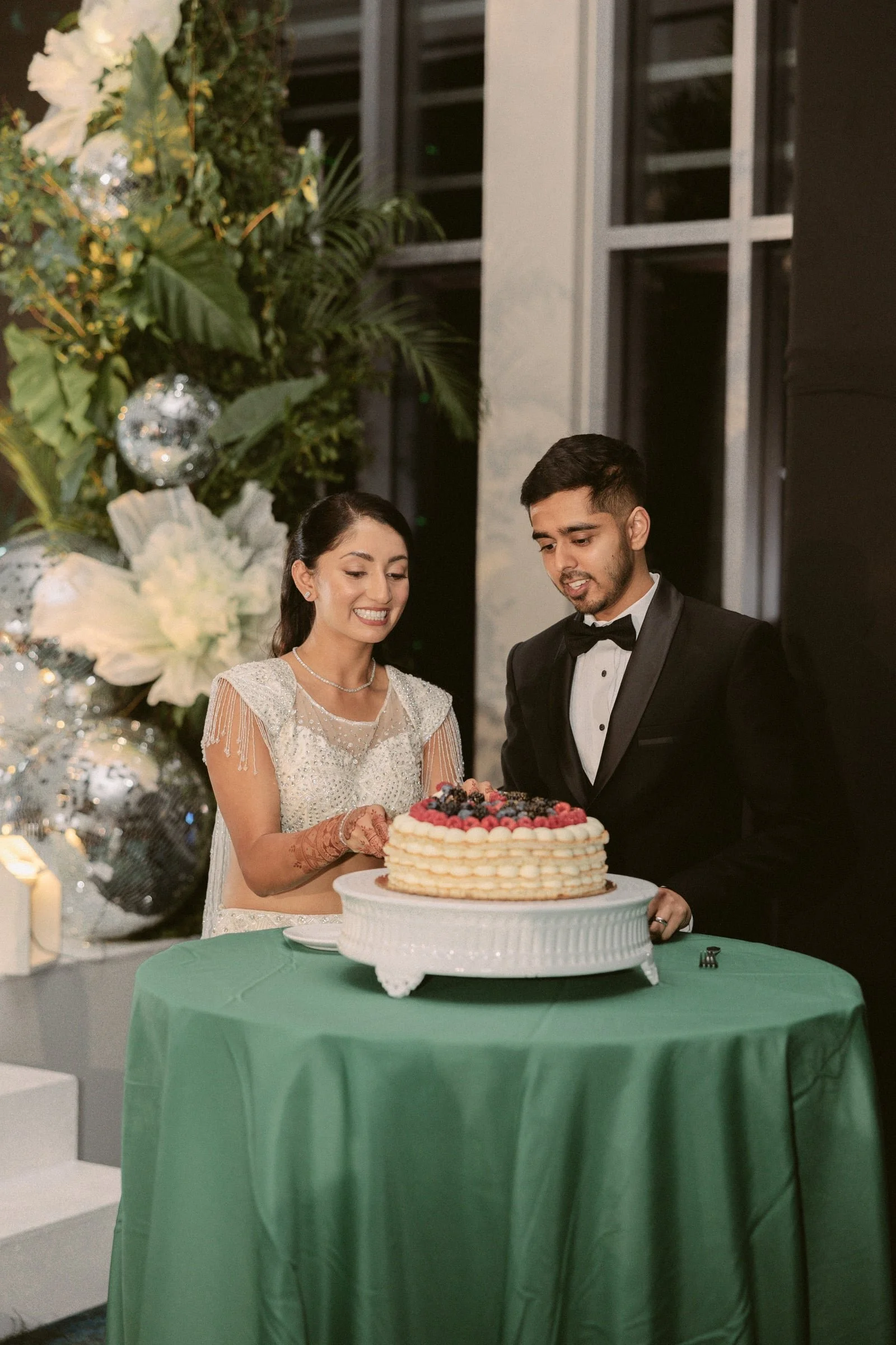 A bride and groom dressed in wedding attire cut a cake at a wedding reception. The bride wears a white dress with beaded details and the groom wears a black tuxedo. They are standing behind a green table with a white cake decorated with berries. Ther