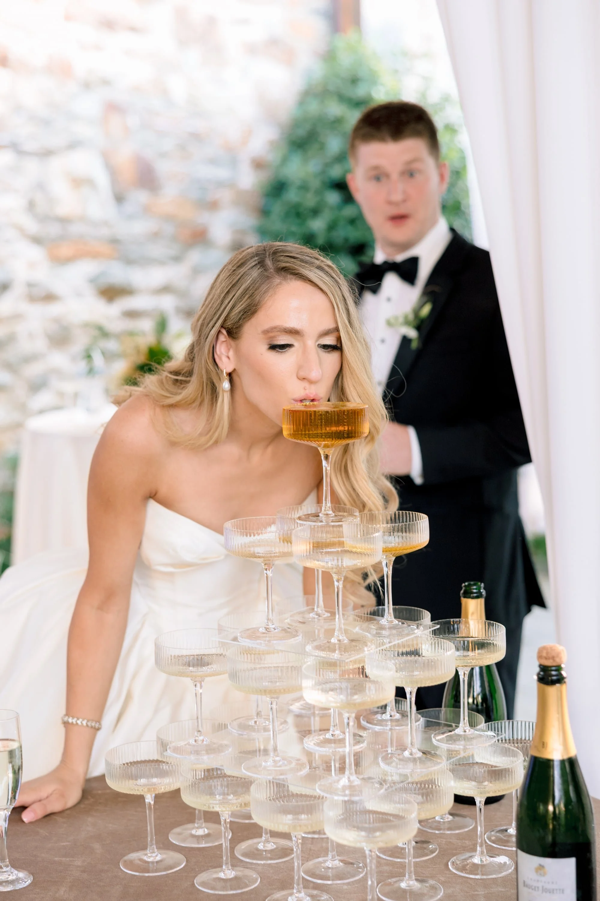 A bride in a white wedding dress and a groom in a tuxedo at a wedding reception, with a tower of champagne glasses and bottles on the table.