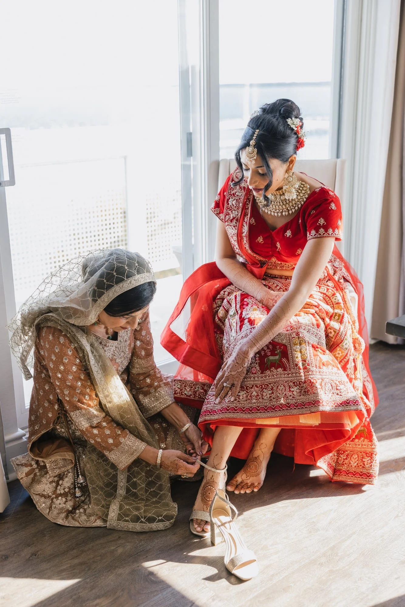 Two women dressed in traditional Indian wedding attire, with one helping the other put on high-heeled sandals, inside a bright room near a large window.