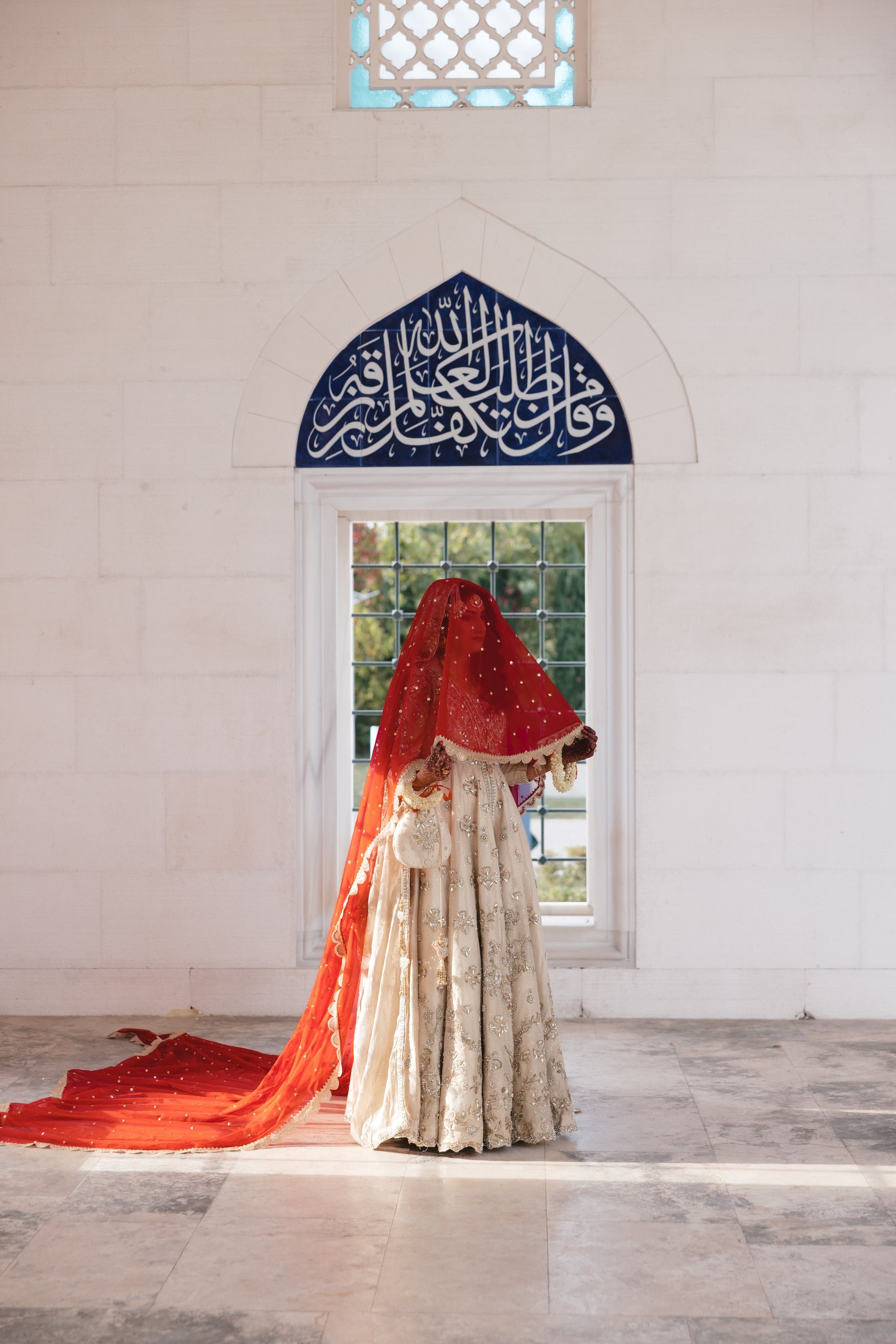 A mannequin dressed in a traditional cream-colored gown with intricate silver embroidery, topped with a red veil that has gold accents, standing in front of a window with a grid pattern, inside a white brick building with a decorative Islamic calligr