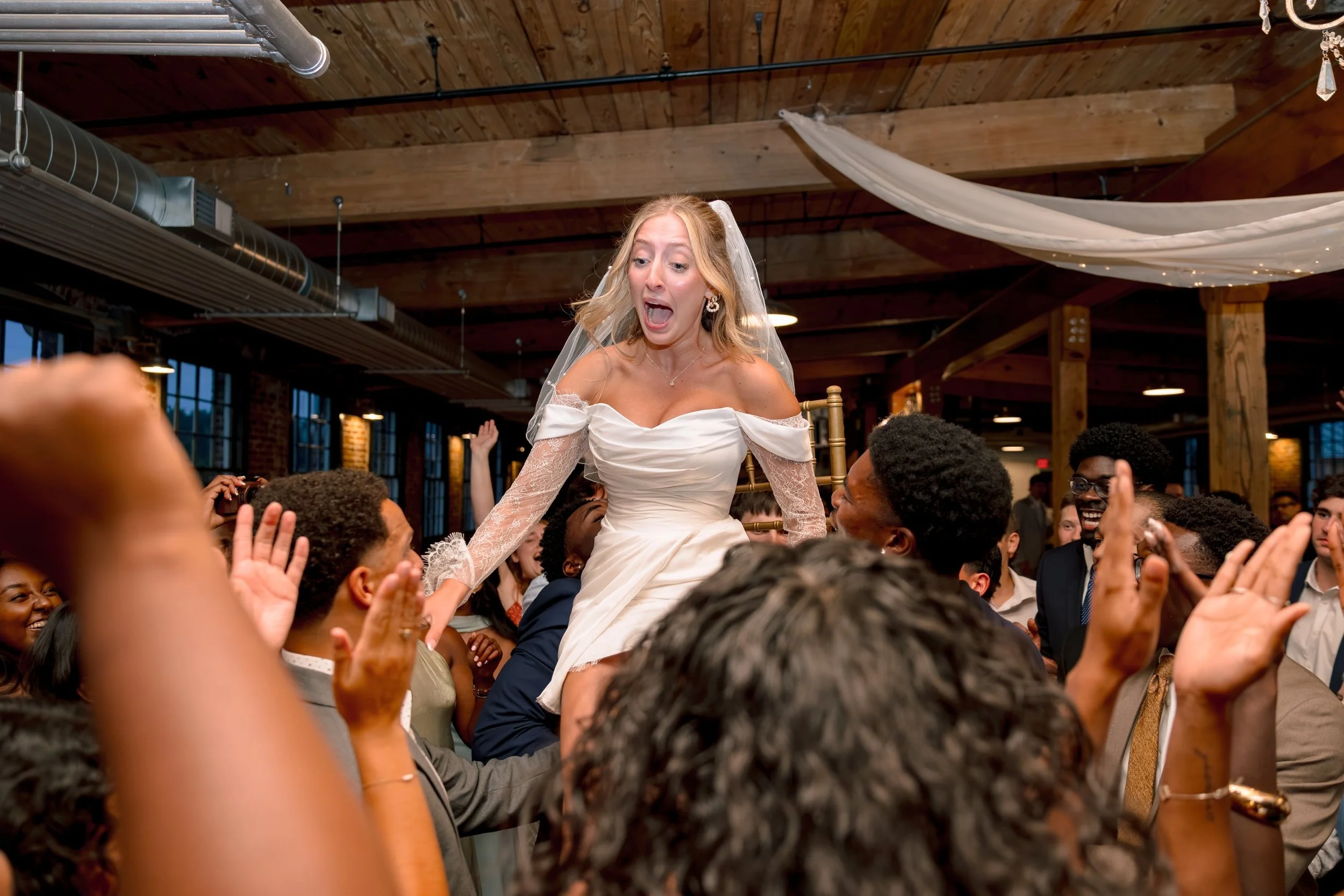 A bride wearing a white wedding dress and veil is being lifted on a chair by a crowd at a wedding celebration, with her mouth open and eyes wide in excitement.