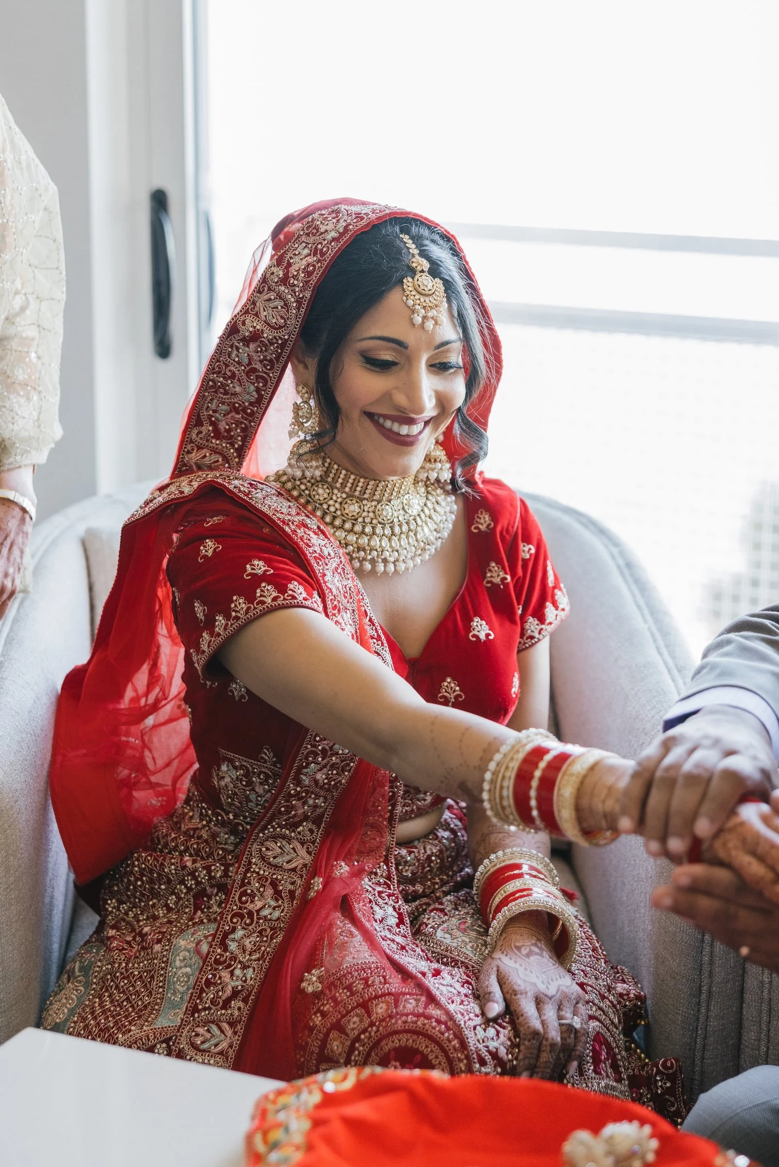 Woman dressed in traditional red Indian wedding attire with gold embroidery, jewelry, and henna, smiling as she is assisted with her wedding bangles.