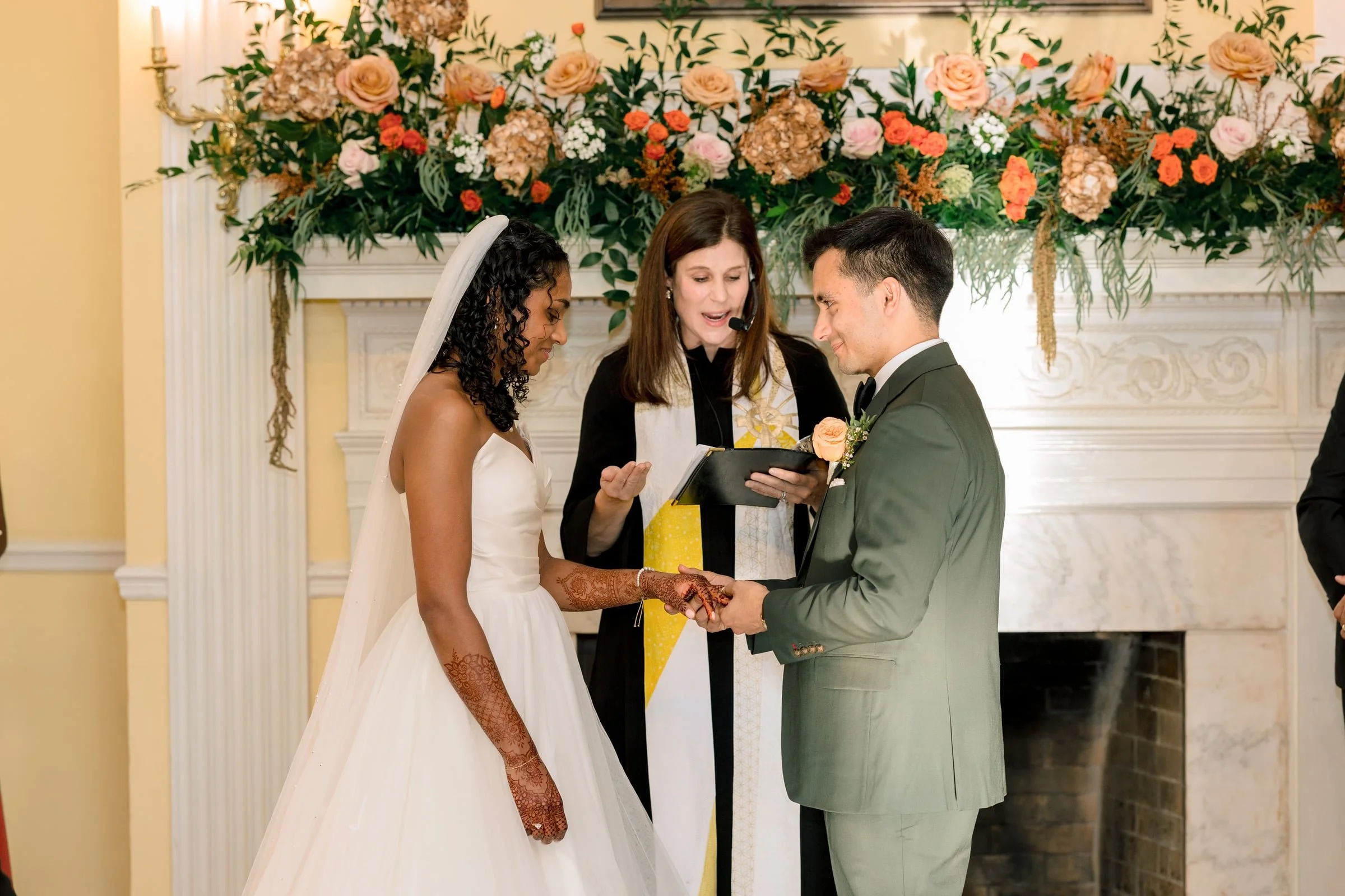 A bride and groom holding hands and looking at each other during their wedding ceremony, with an officiant standing between them, against a backdrop of large floral arrangements.