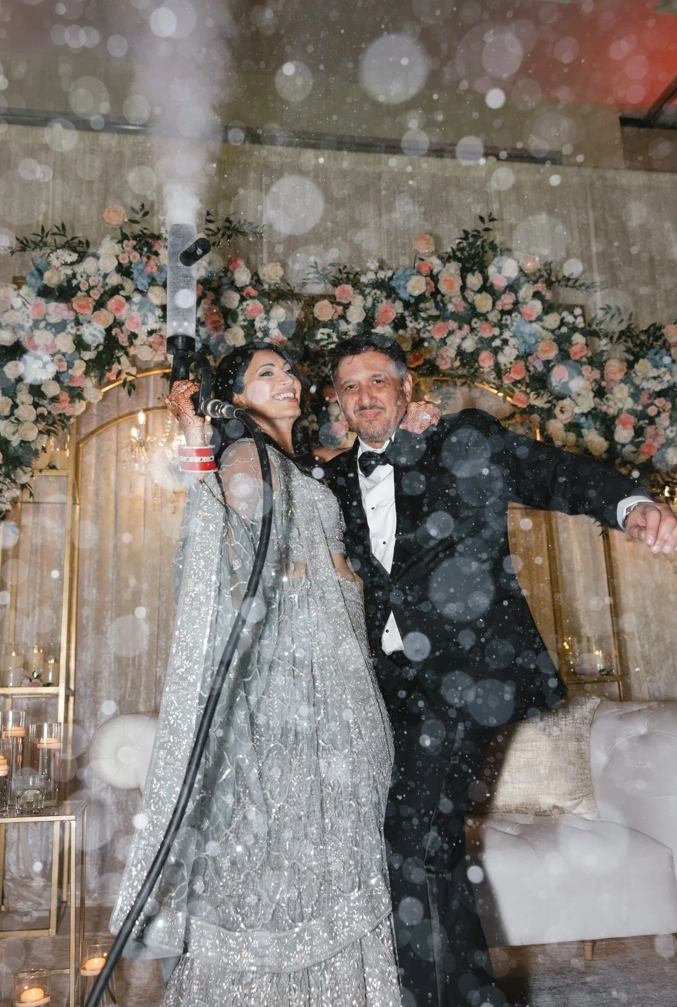 A bride and groom celebrating at their wedding reception, with floral decorations in the background, as the bride sprays champagne.